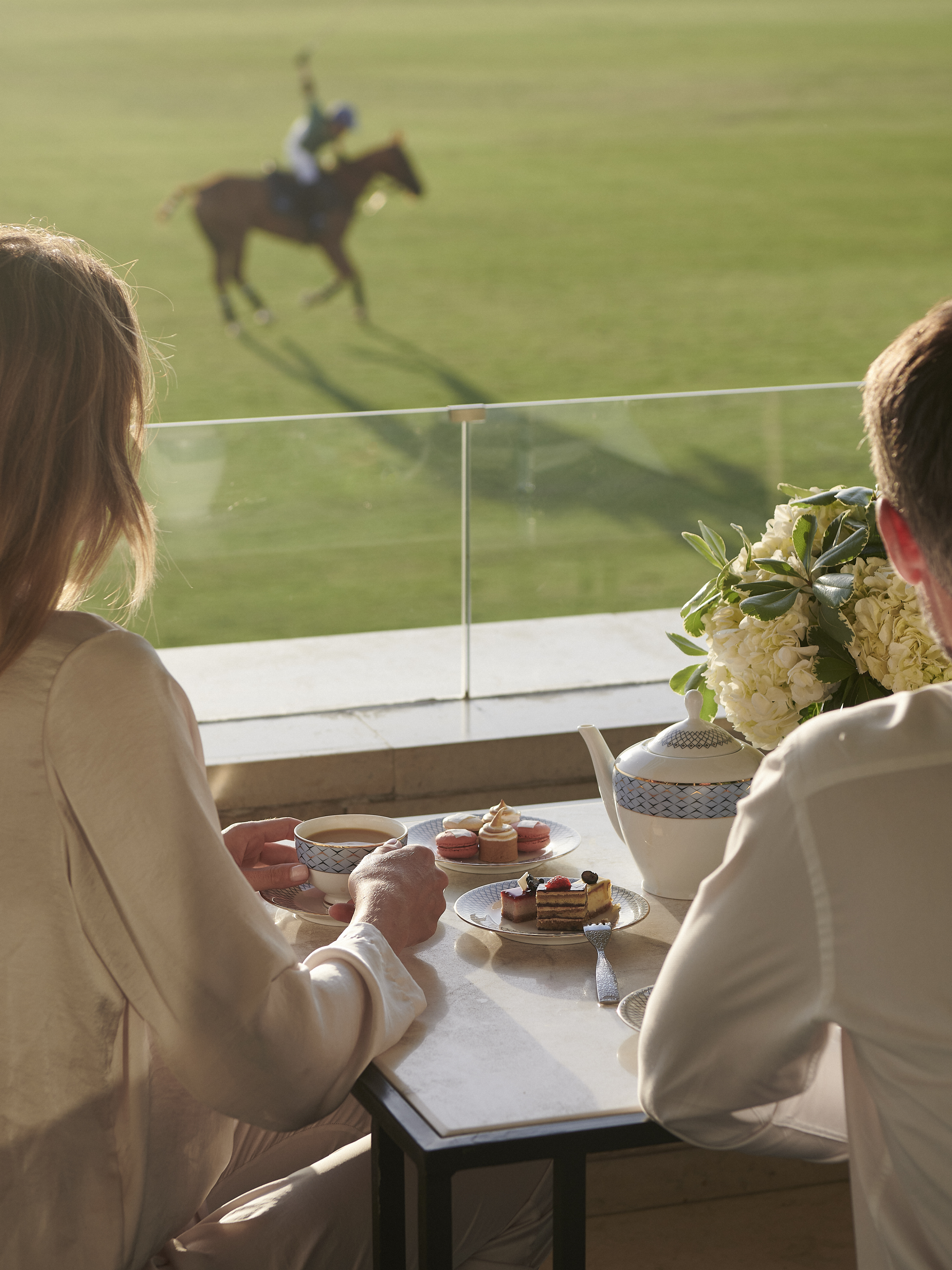 Couple enjoying afternoon tea and desserts on a balcony, watching a polo match.