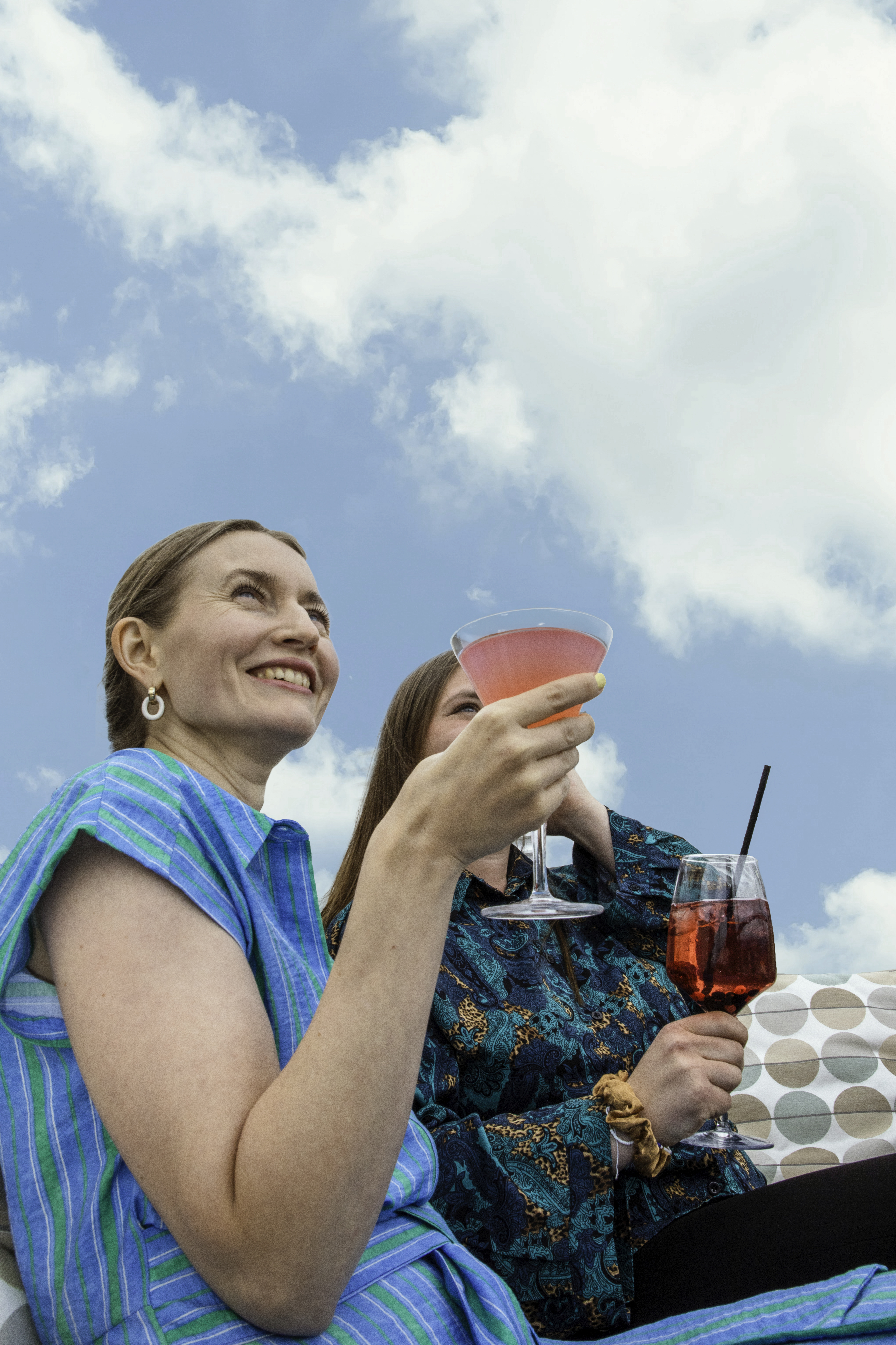 Smiling women raising colorful cocktails against a vibrant blue sky with clouds.