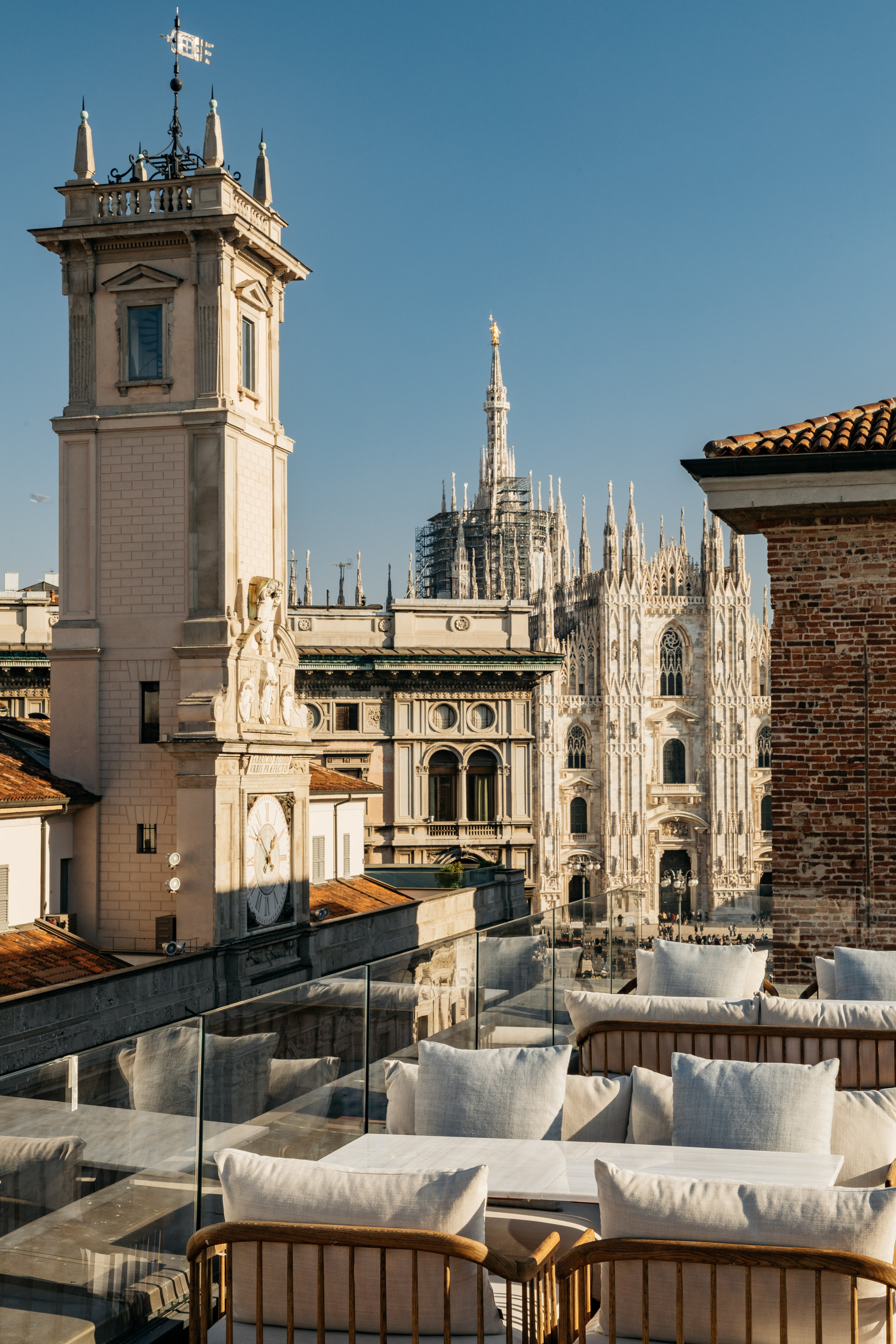 Rooftop terrace view of Milan Duomo cathedral and historic clock tower.