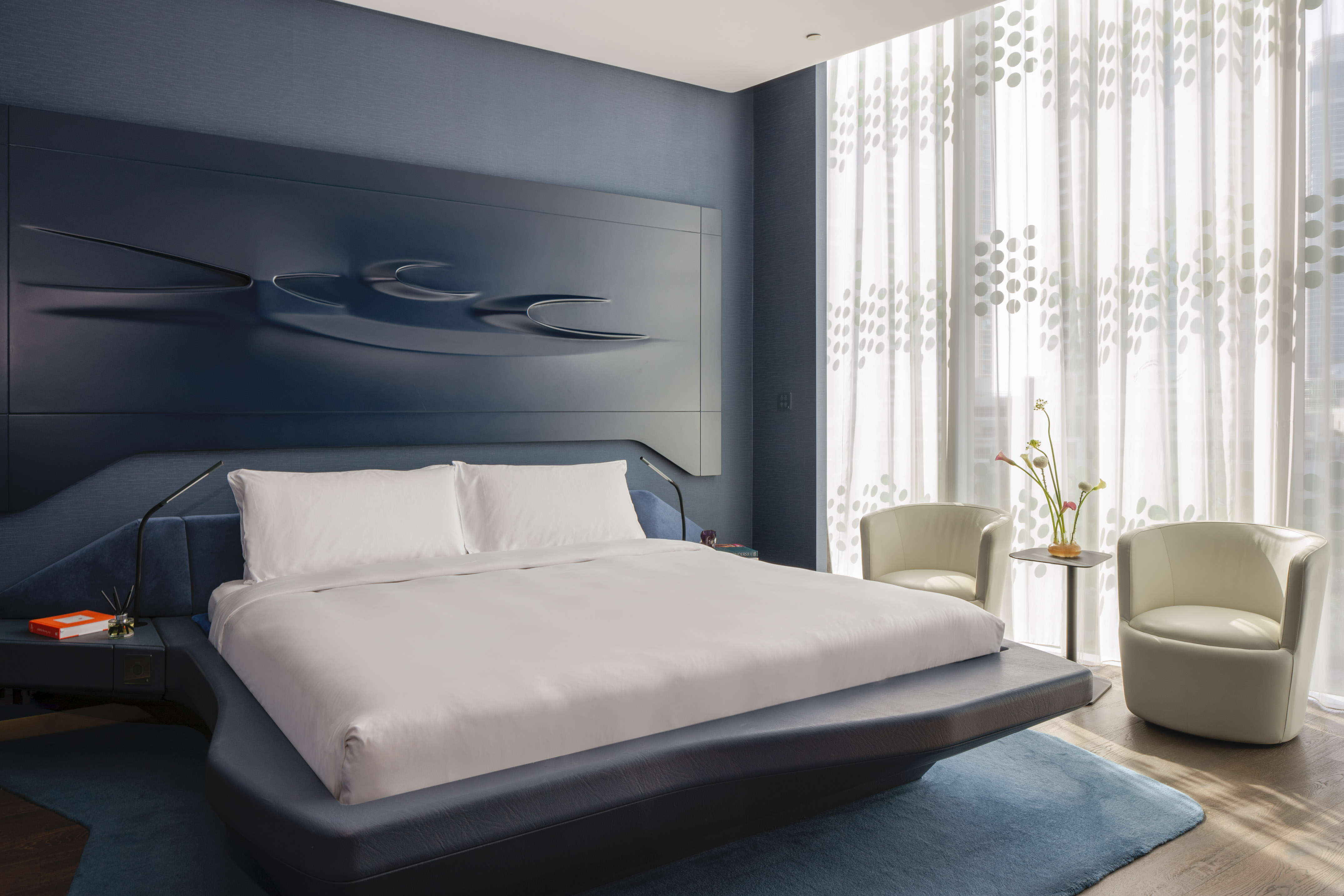 Modern blue bedroom with king bed, sculpted headboard, and sheer curtains.