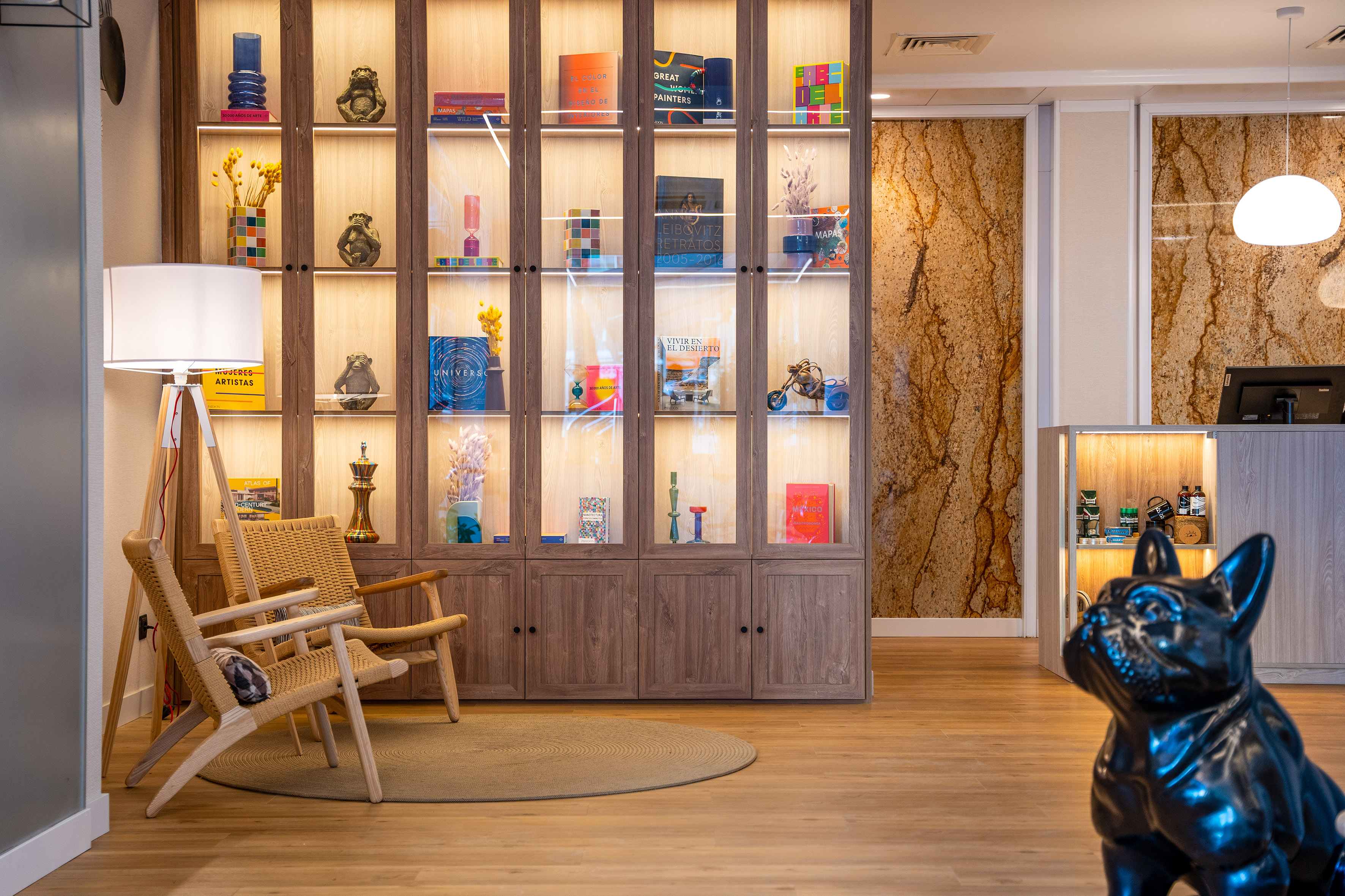 Modern hotel reception with illuminated display cabinet, woven chairs, and bulldog decor.