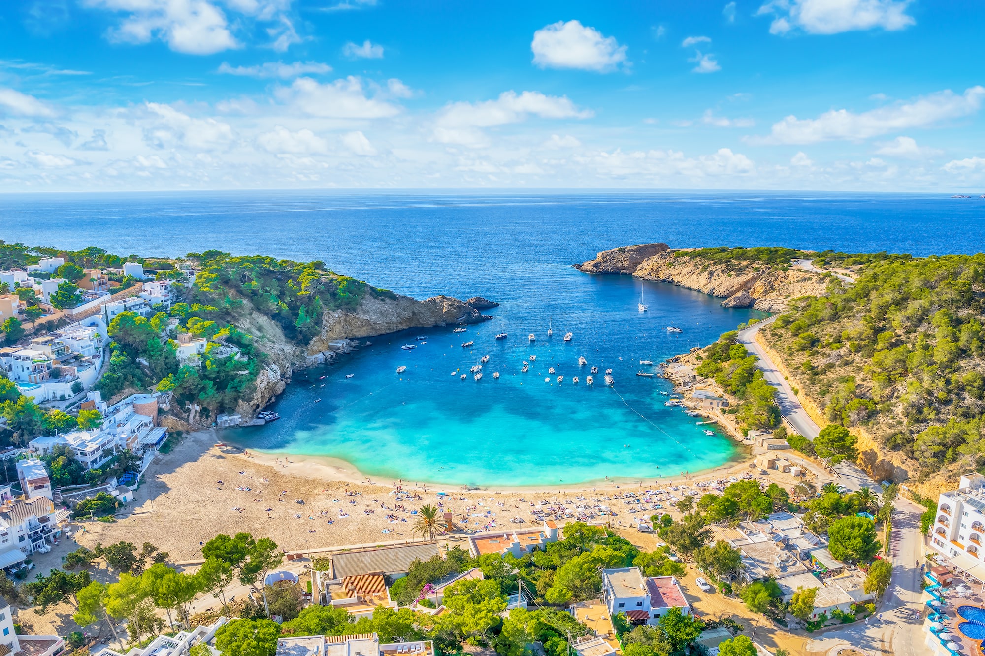 Cala mediterránea con playa de arena, mar turquesa, barcos y casas.