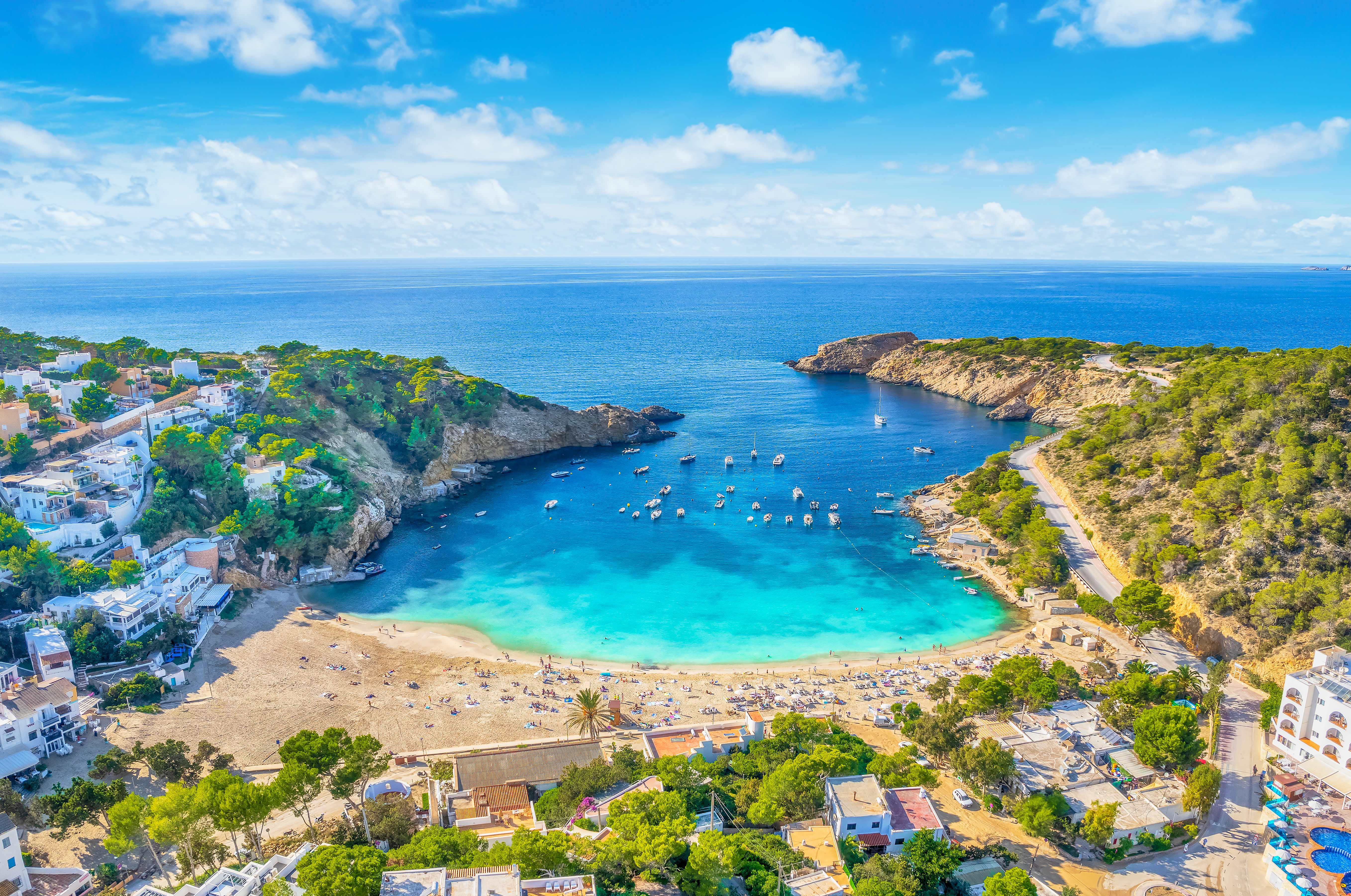 Cala mediterránea con playa de arena, mar turquesa, barcos y casas.