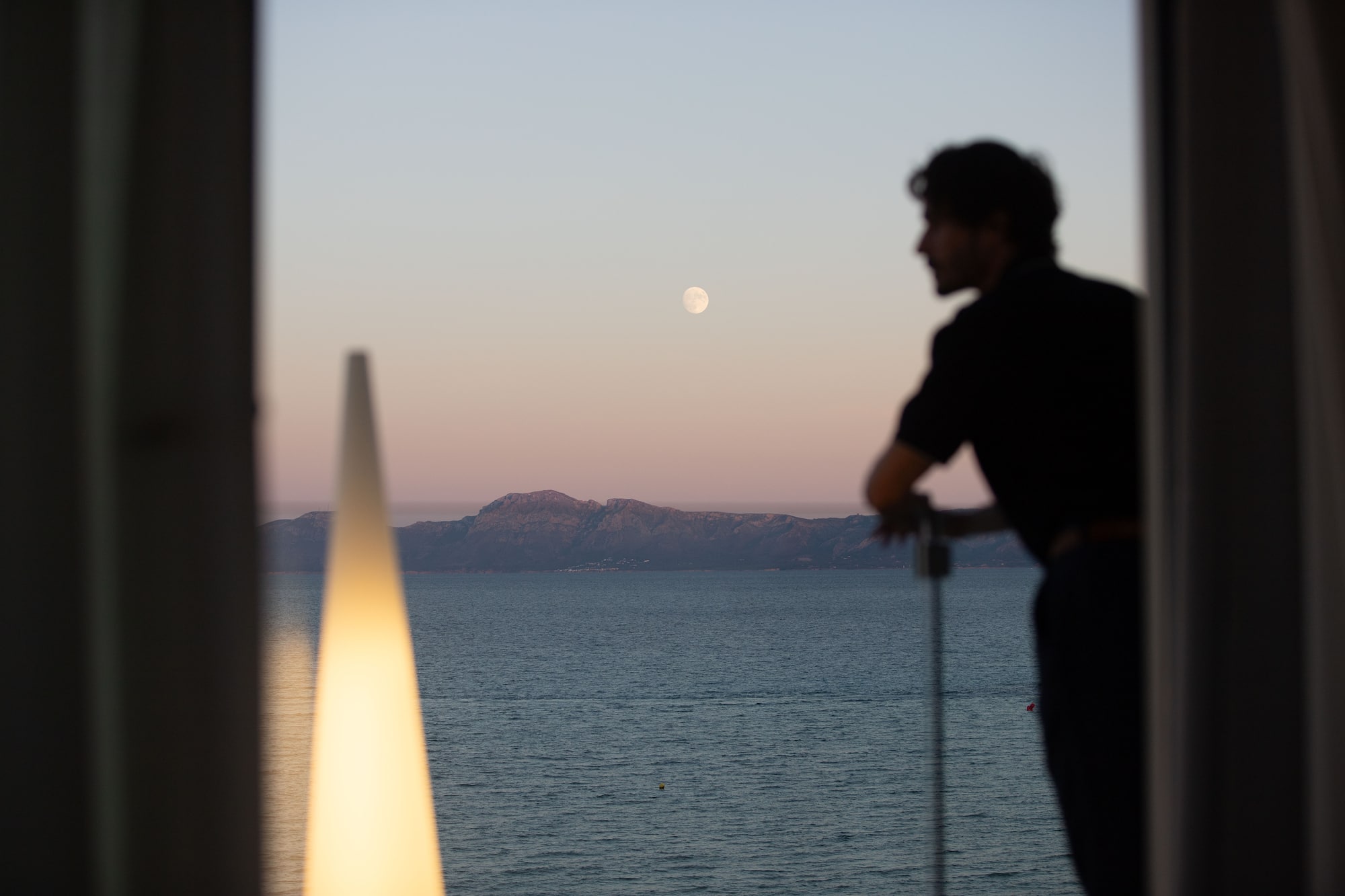 Man in silhouette on balcony watching full moon over ocean and mountains.