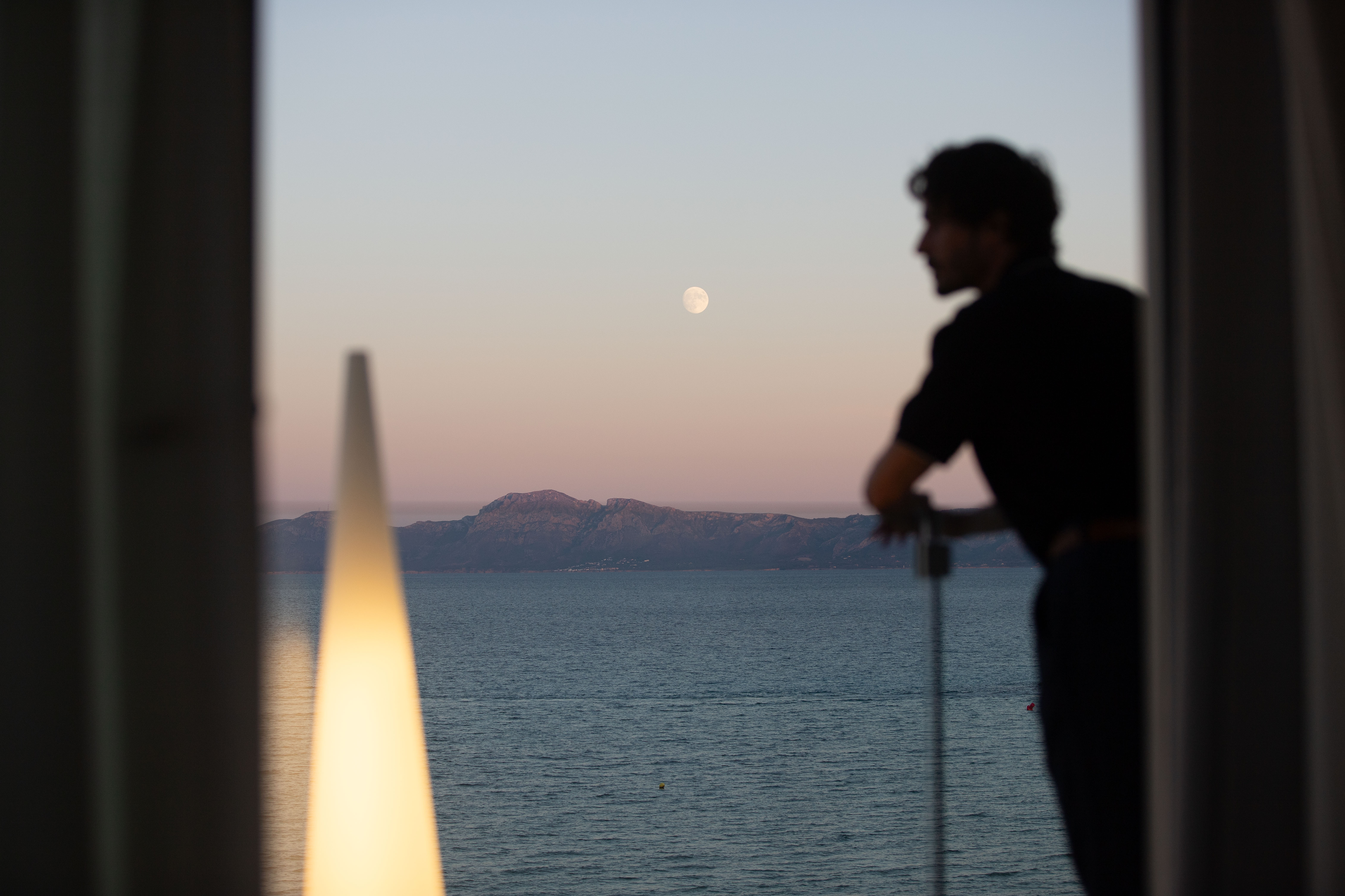 Man in silhouette on balcony watching full moon over ocean and mountains.