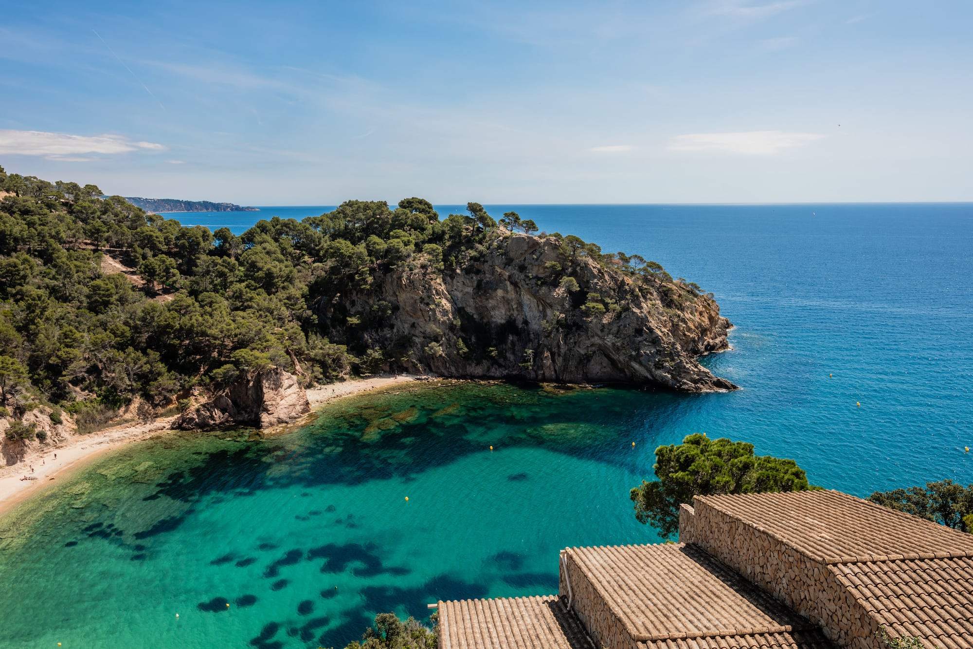 Turquoise bay with sandy beach, pine-covered cliffs, and terracotta roofs.