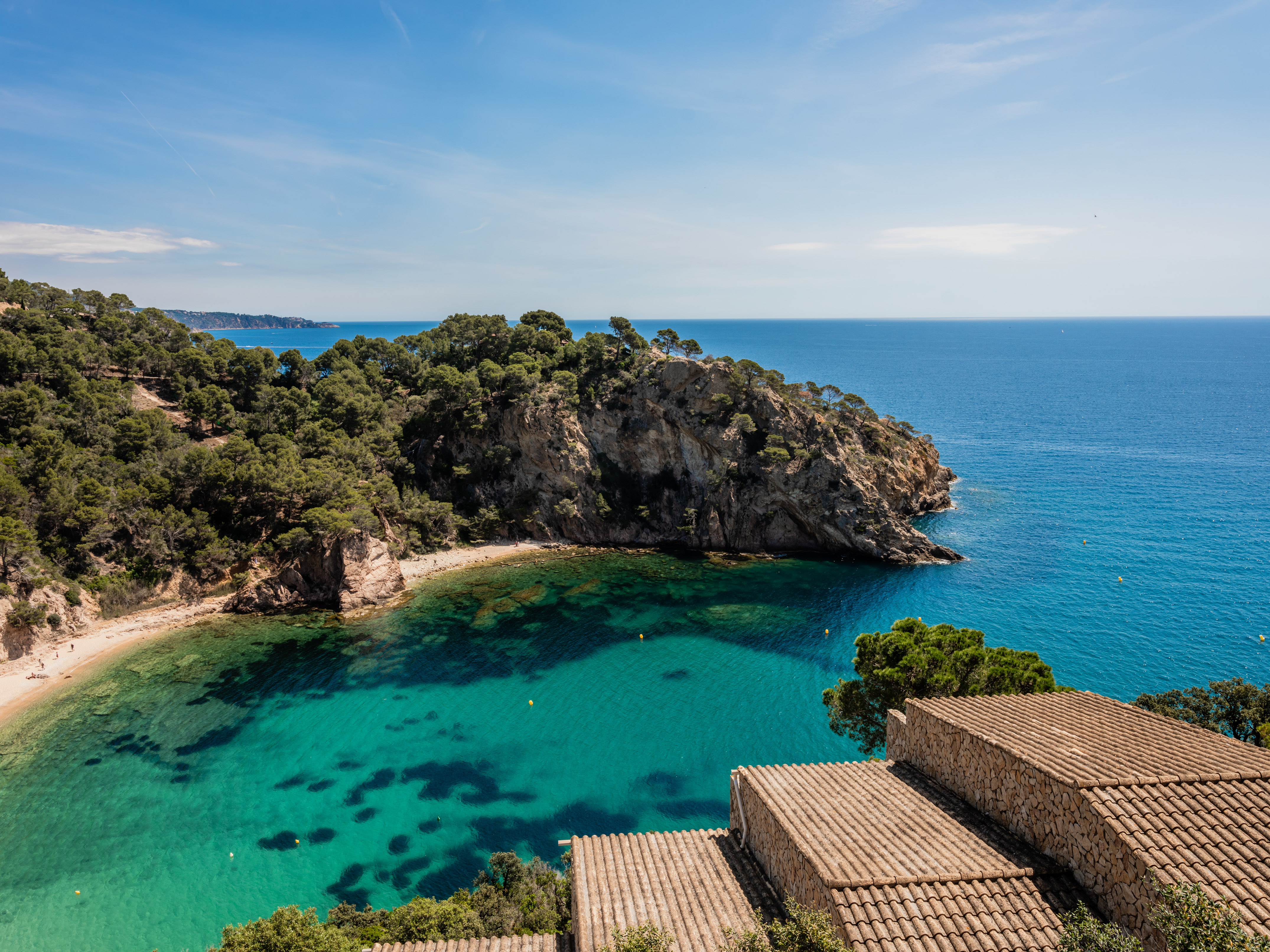 Turquoise bay with sandy beach, pine-covered cliffs, and terracotta roofs.