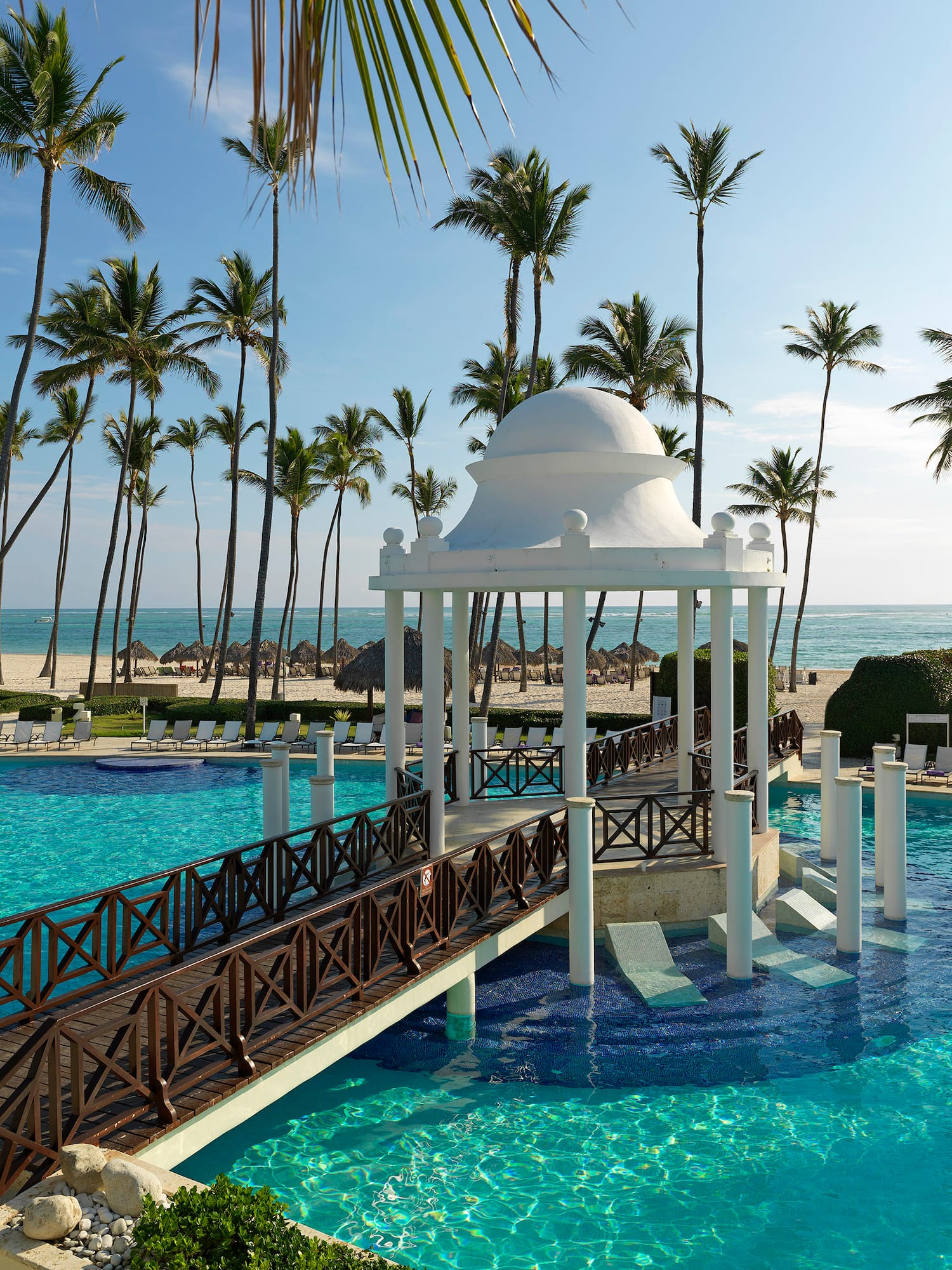 Tropical resort pool, white gazebo, wooden bridge, palm trees, beach view.
