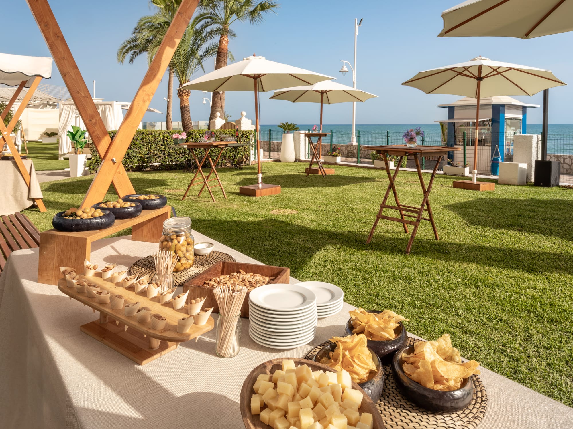 Buffet de catering exterior con aperitivos, jardín, palmeras y mar de fondo.