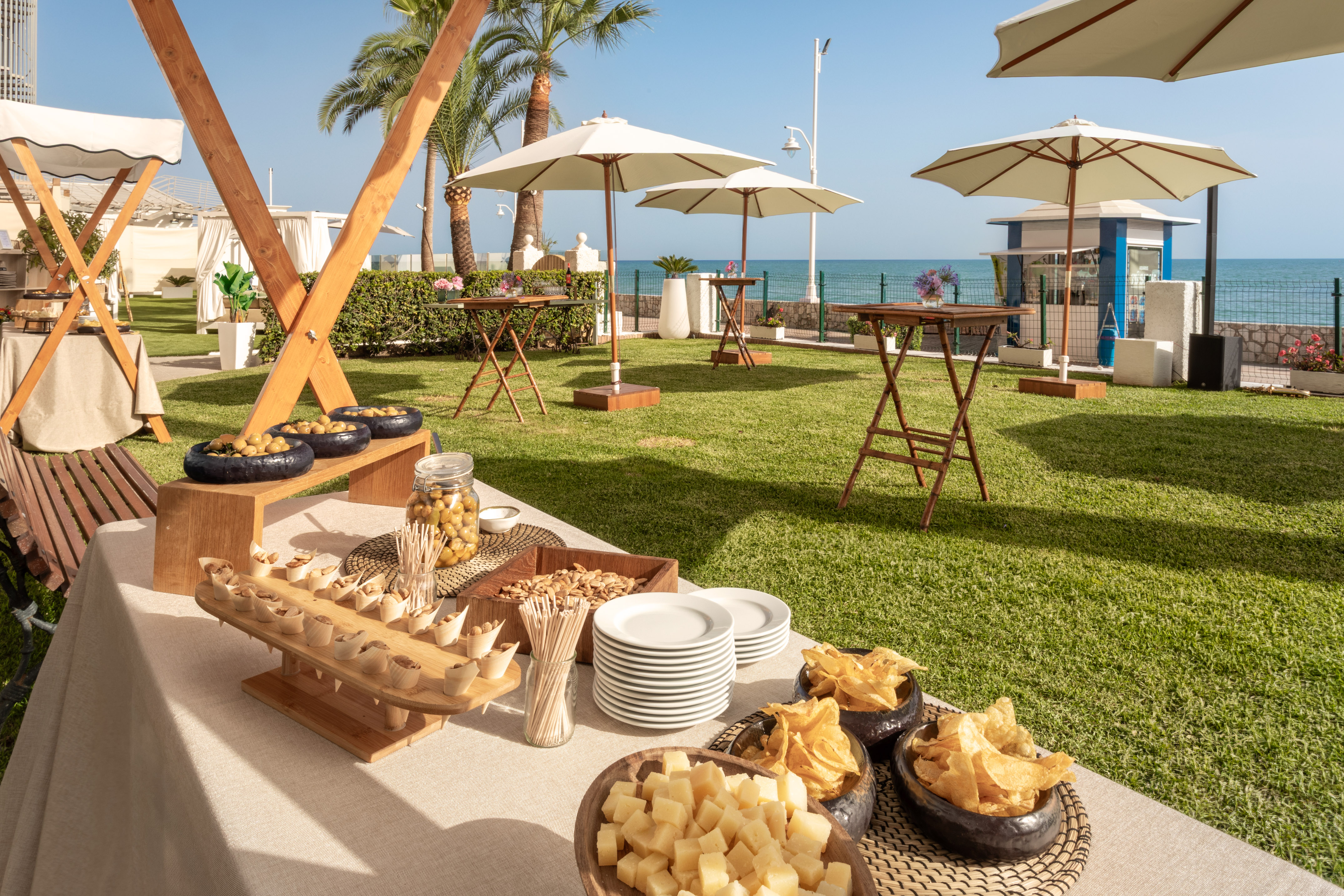 Buffet de catering exterior con aperitivos, jardín, palmeras y mar de fondo.