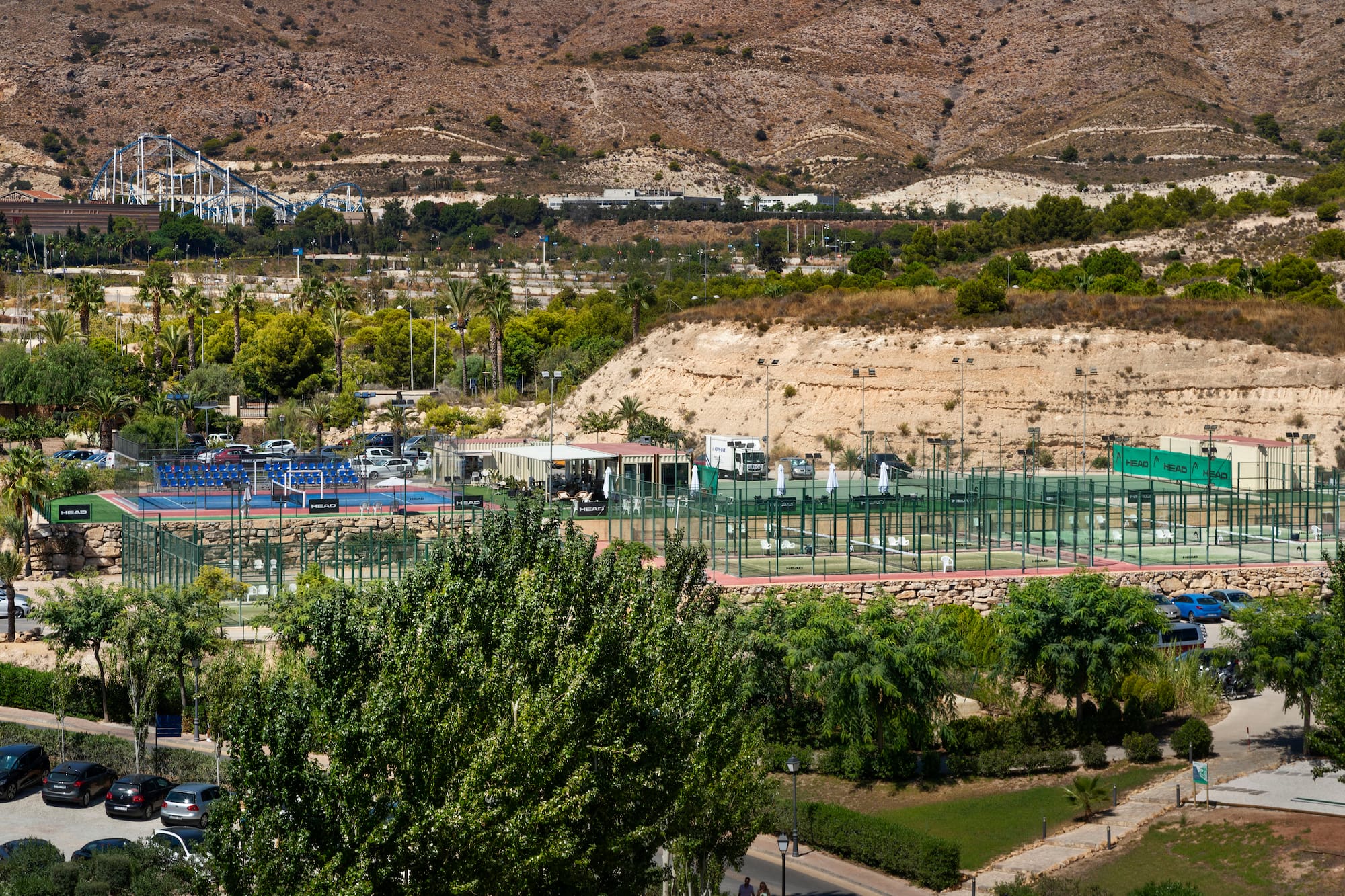 Outdoor padel courts with green trees, dry mountains, and a distant roller coaster.