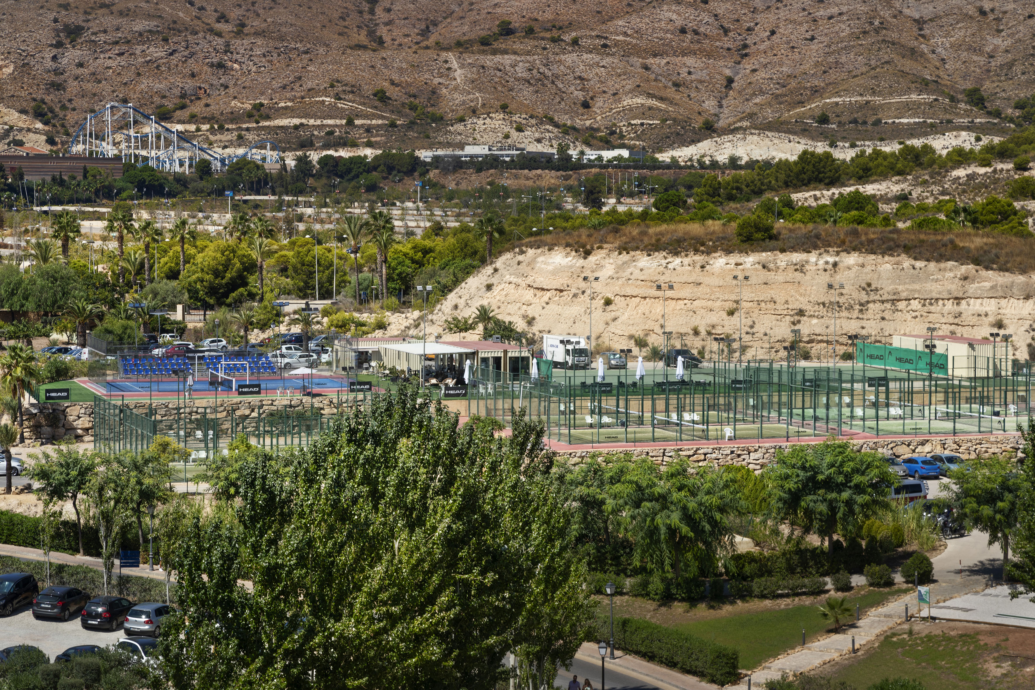 Outdoor padel courts with green trees, dry mountains, and a distant roller coaster.