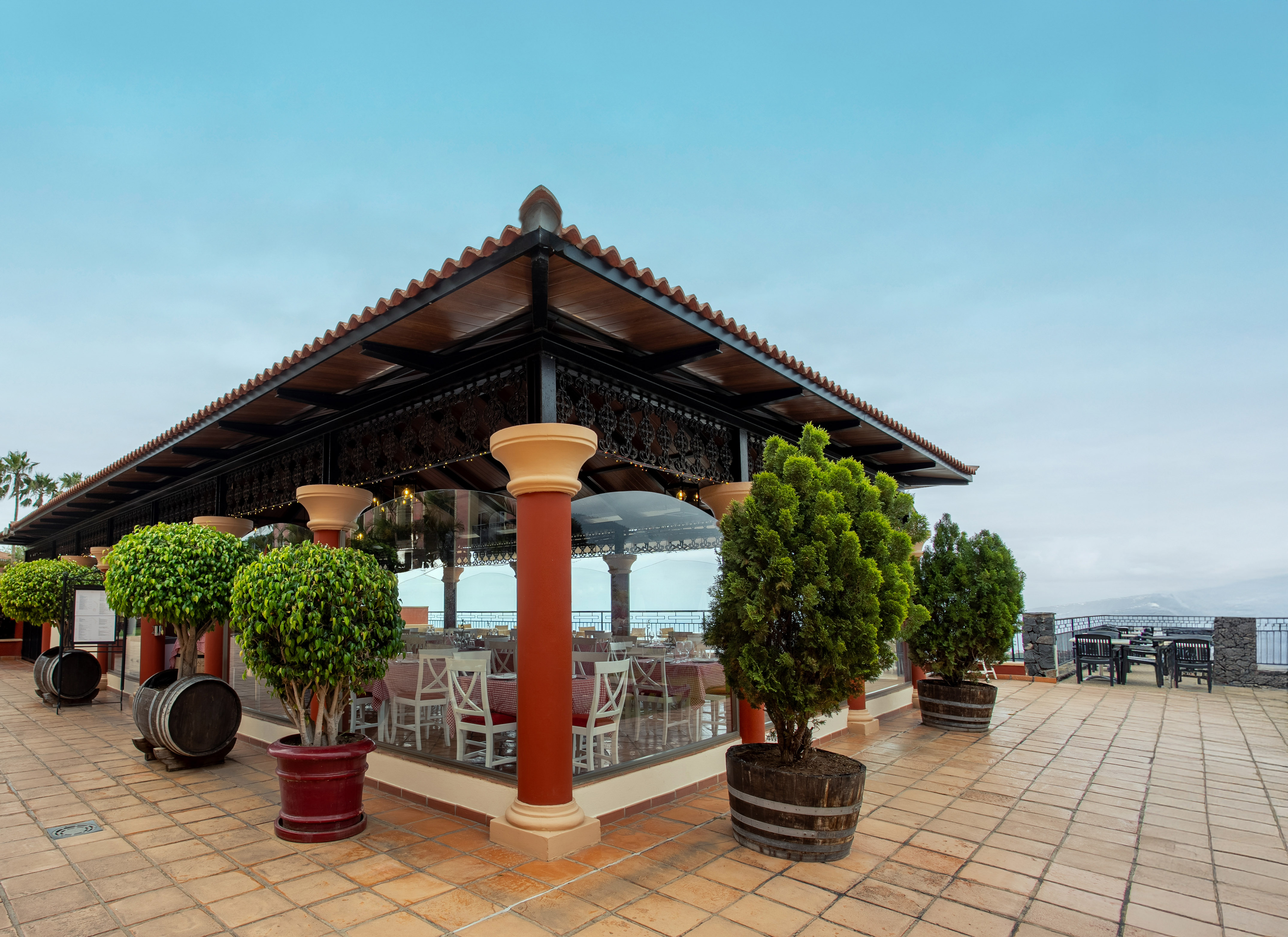 Restaurant terrace with red tiled roof, glass walls, and lush potted greenery.