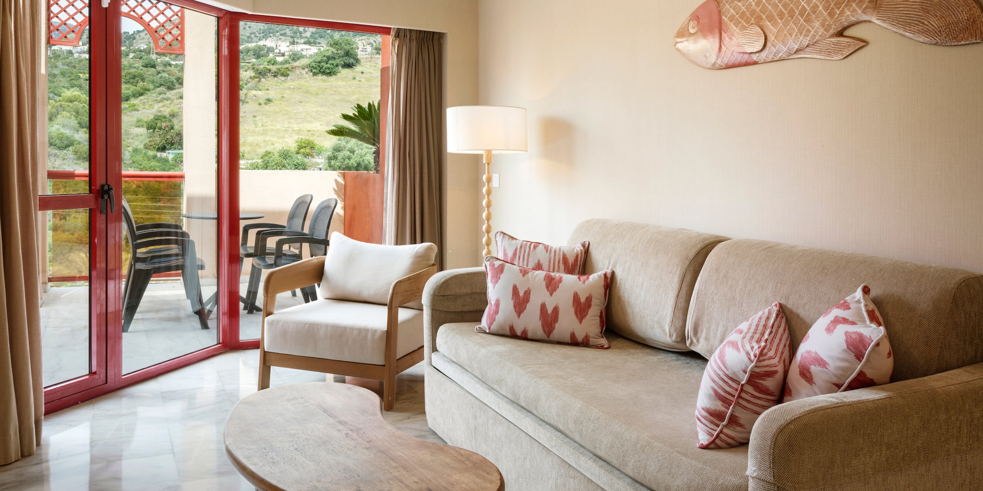 Hotel living room with sofa, armchair, and balcony with green hill view.