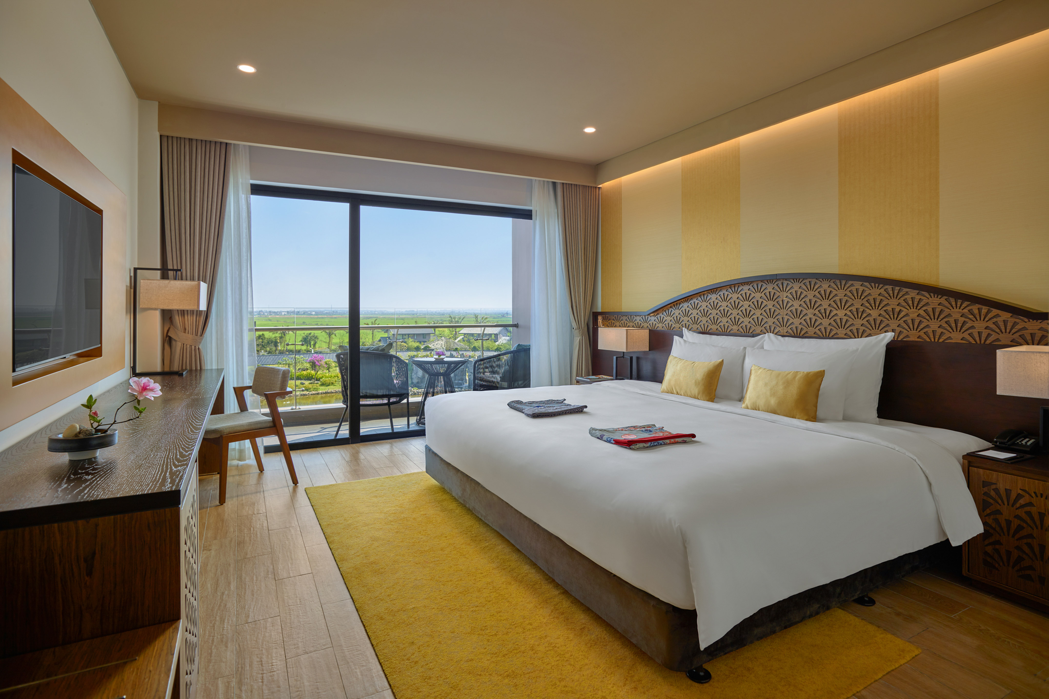 Modern hotel room with king bed, yellow decor, and balcony view of green landscape.