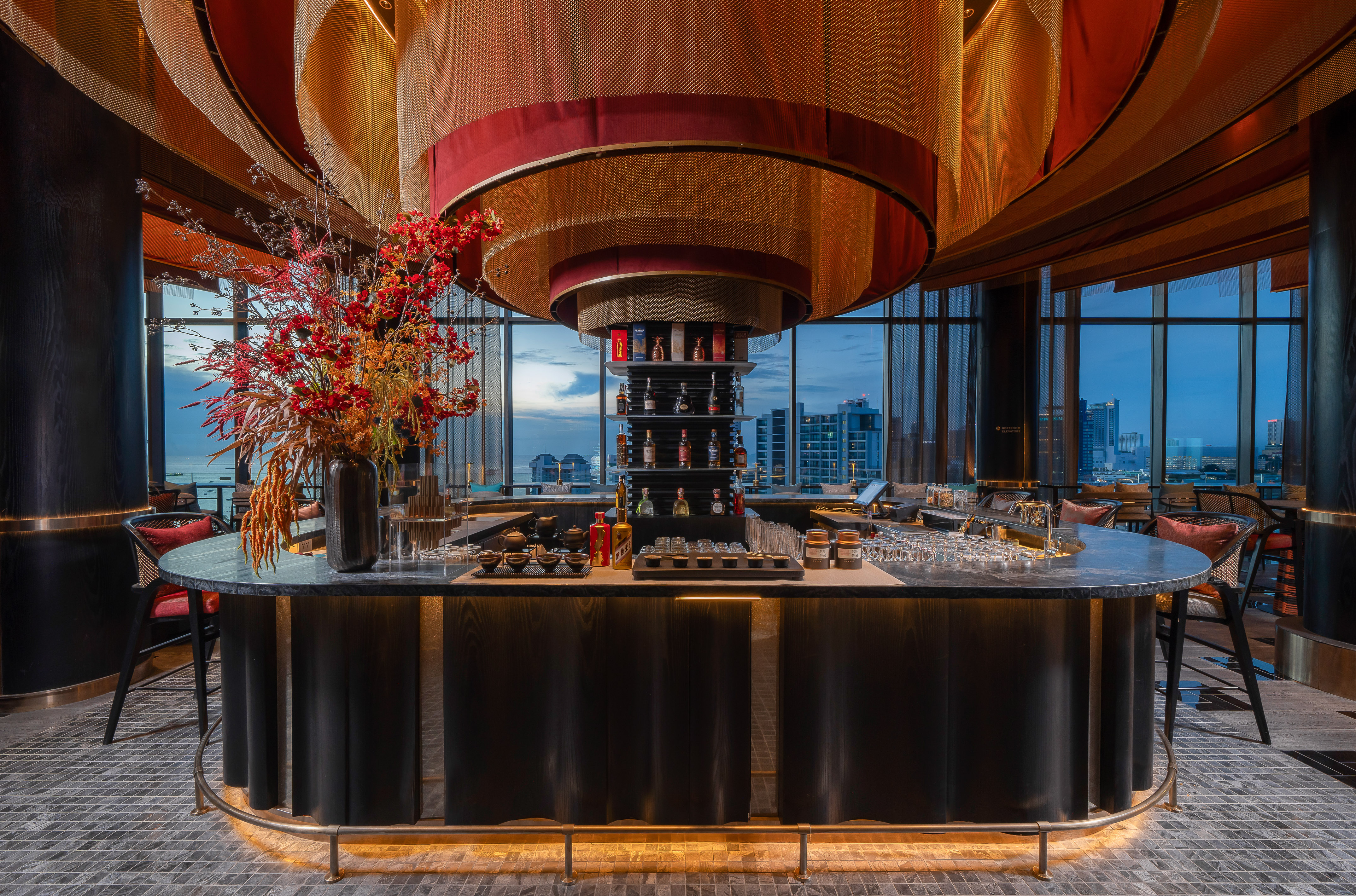 Luxury rooftop bar with circular counter, liquor display, floral decor, cityscape.