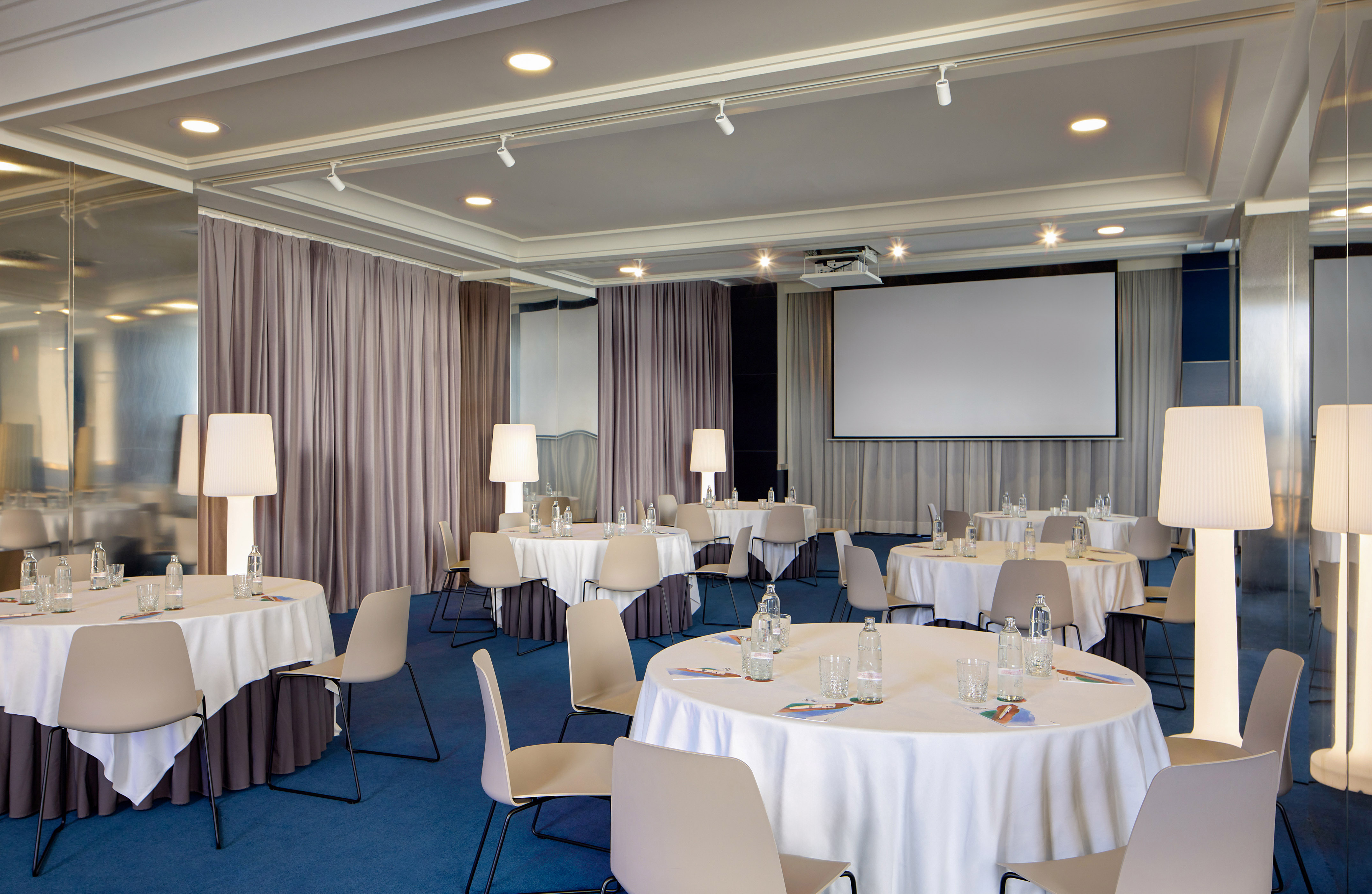 Spacious meeting room with round tables, chairs, projector screen, and blue carpet.