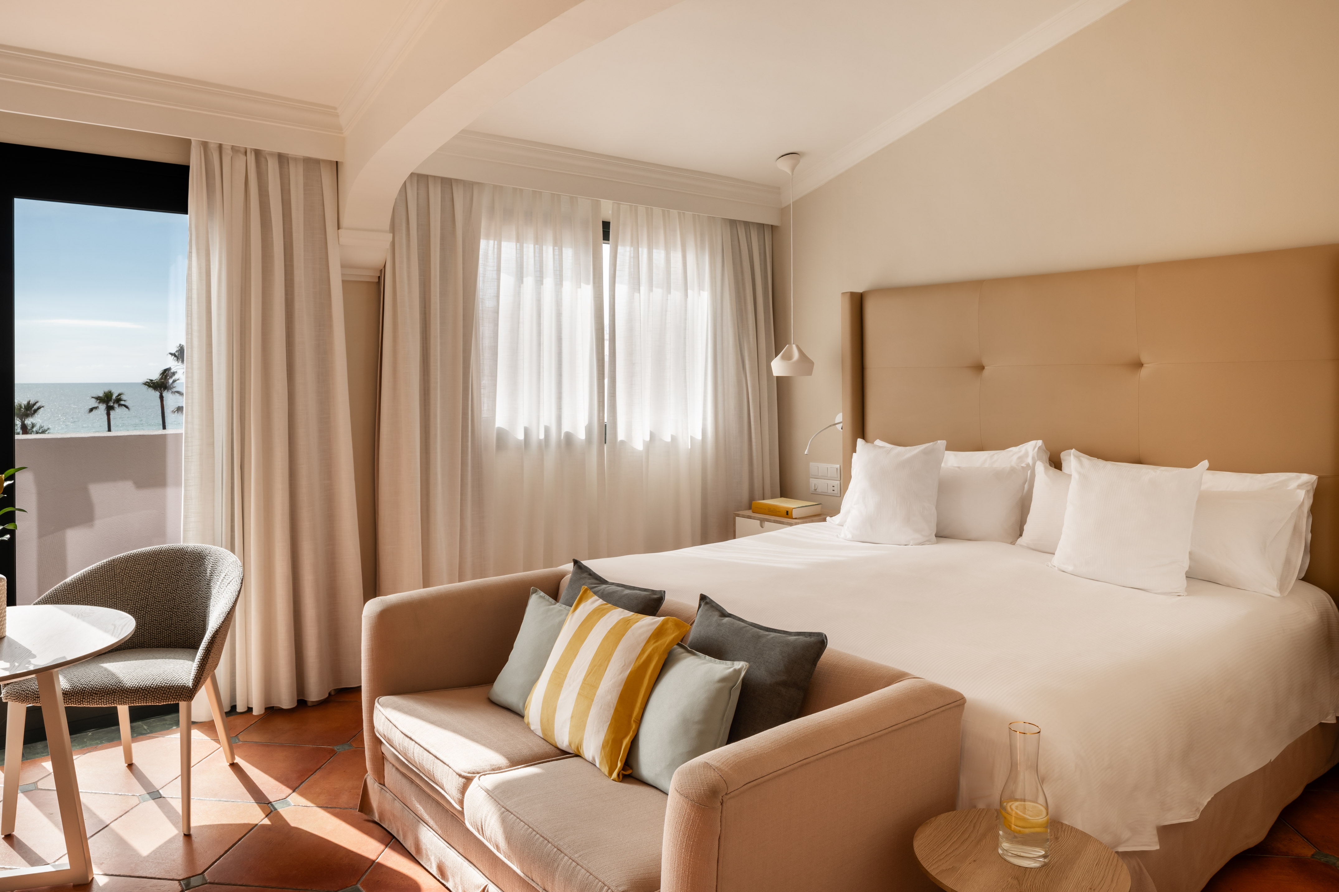 Hotel room with ocean view balcony, large bed, and sofa with accent pillows.