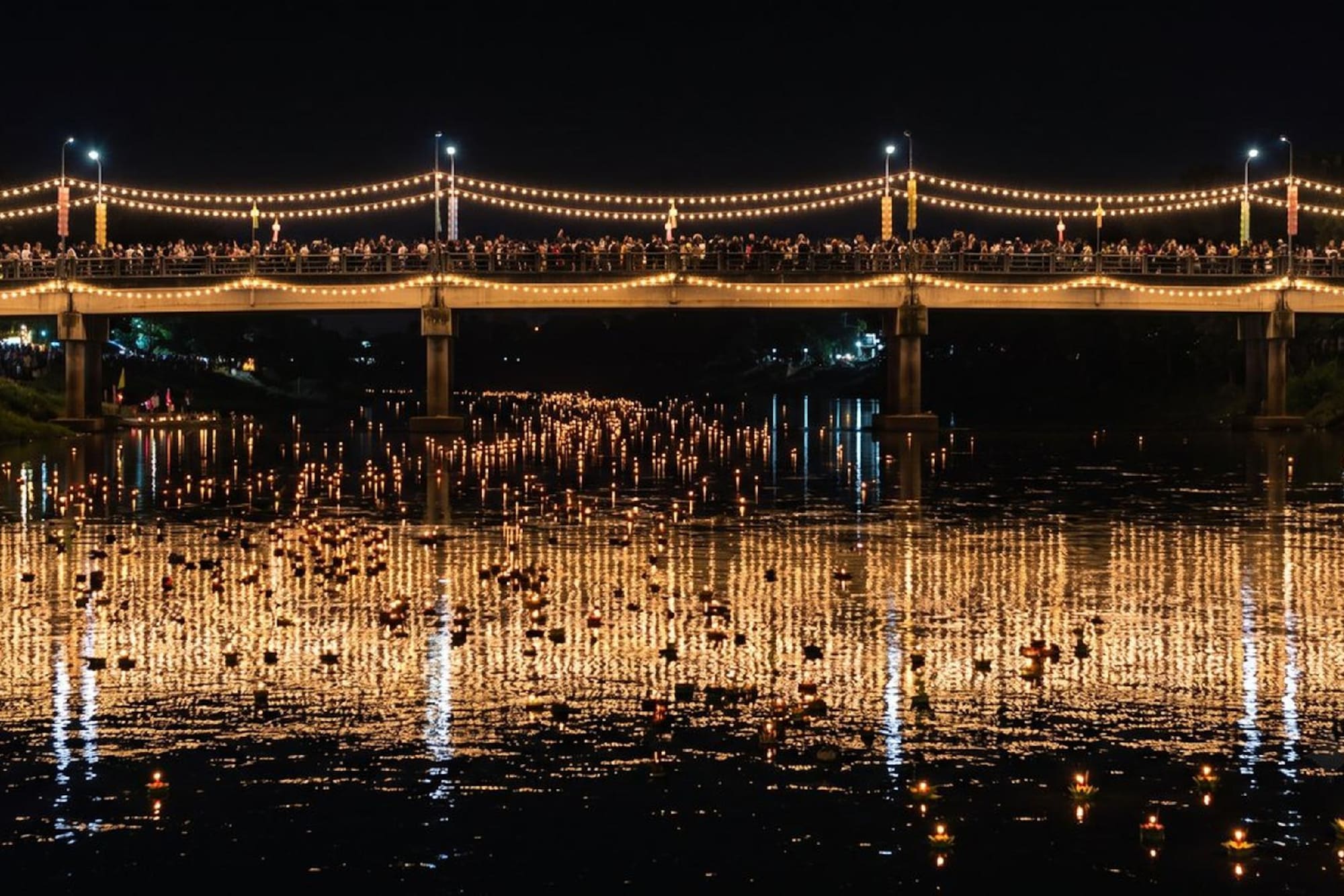 Loy Krathong festival at night, with glowing krathongs on river and lit bridge.