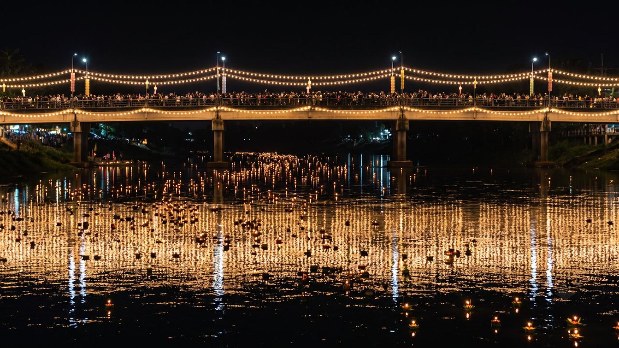 Loy Krathong festival at night, with glowing krathongs on river and lit bridge.