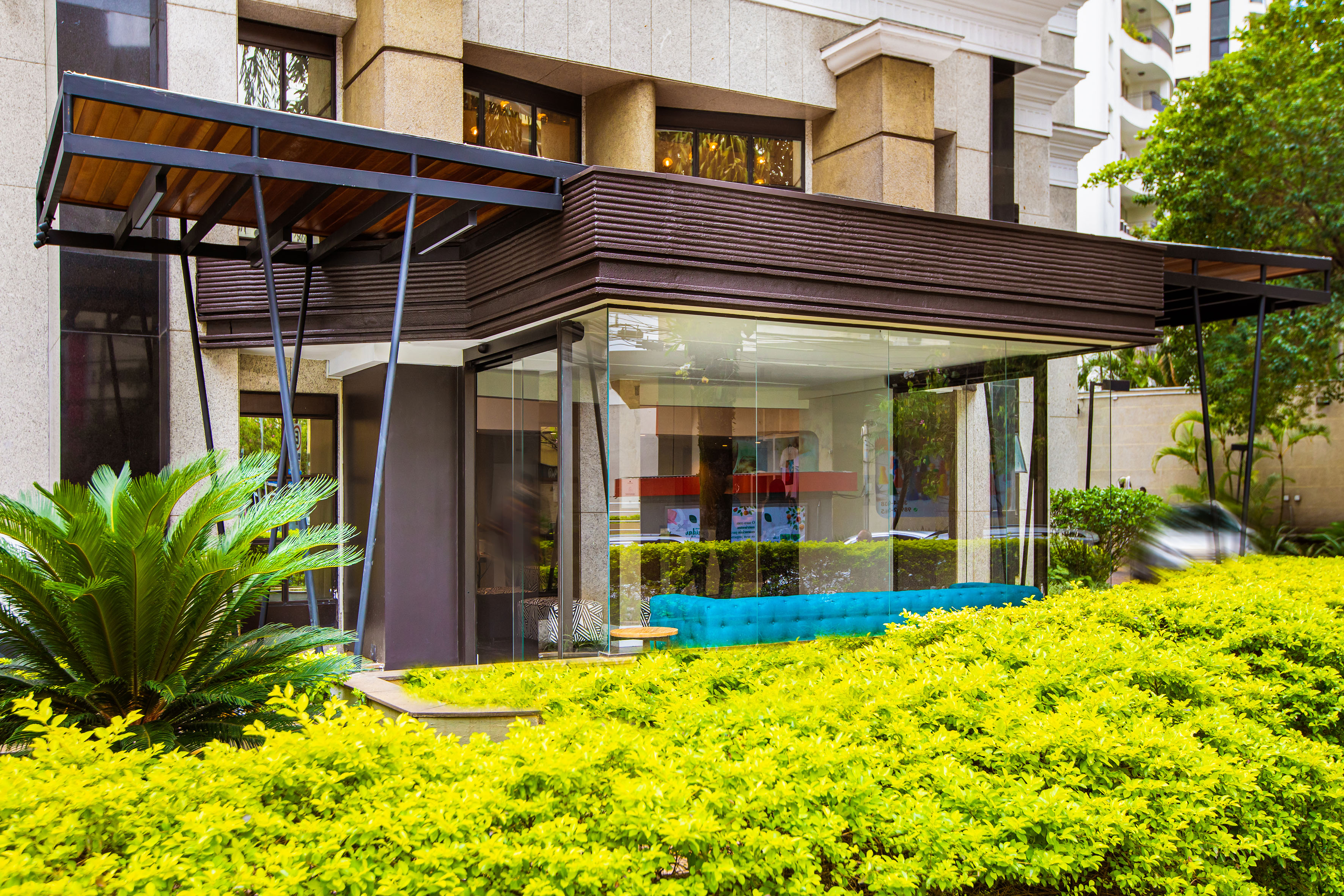 Modern building entrance with glass, wood canopy, teal sofa, and lush landscaping.