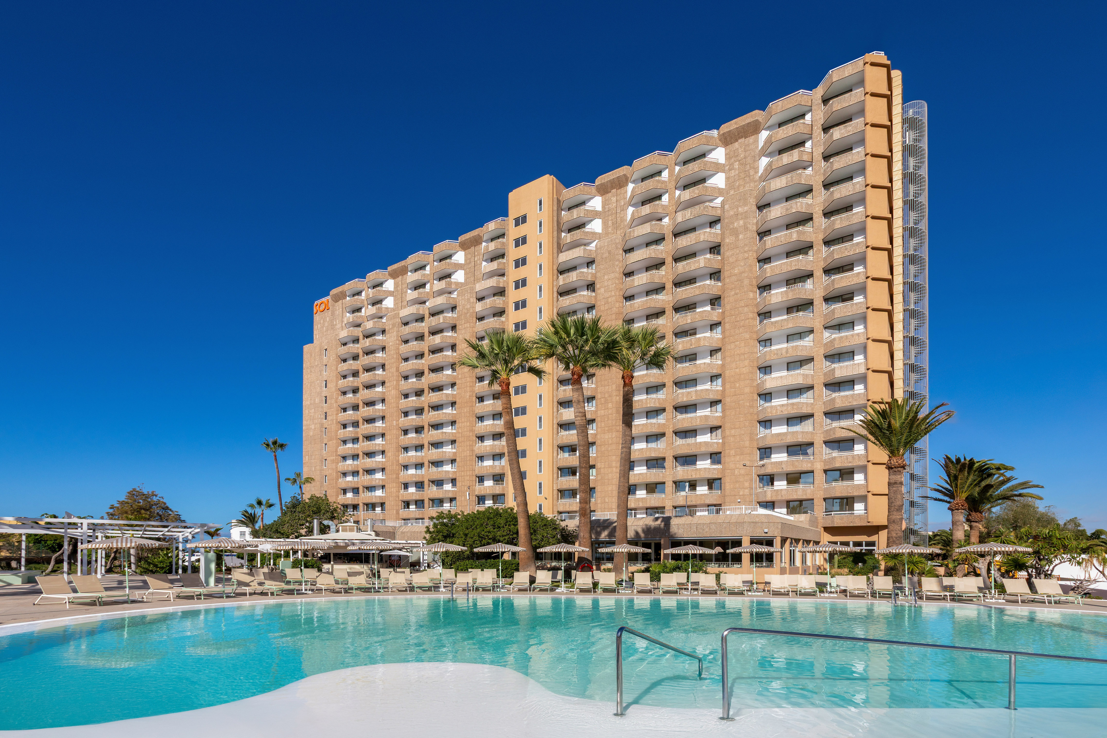 Sol hotel resort with large swimming pool, palm trees, and balconies under blue sky.