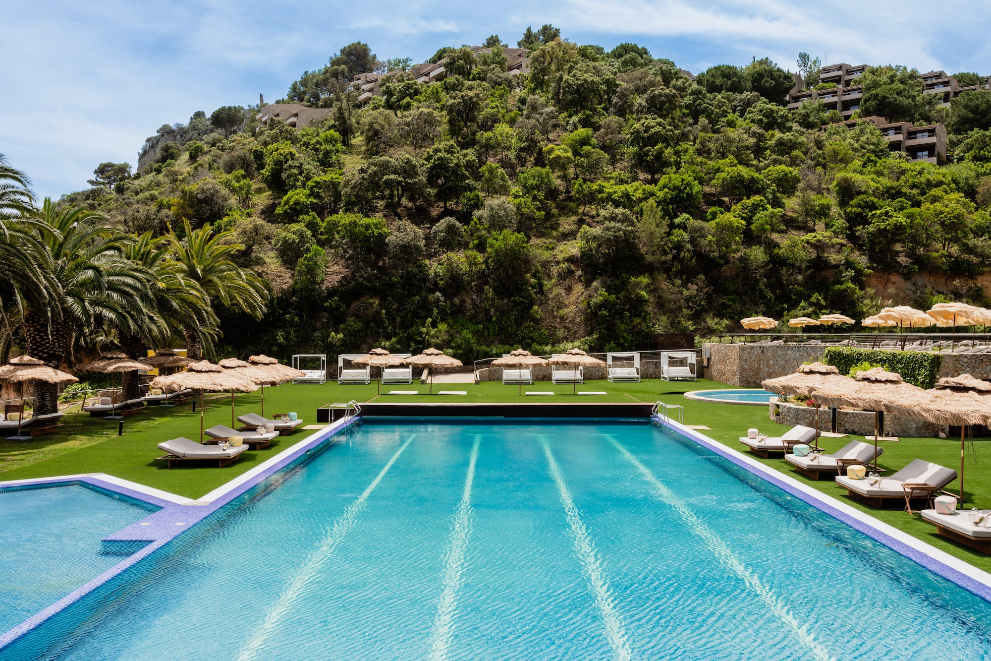 Resort swimming pool with sun loungers, straw umbrellas, and lush hillside.