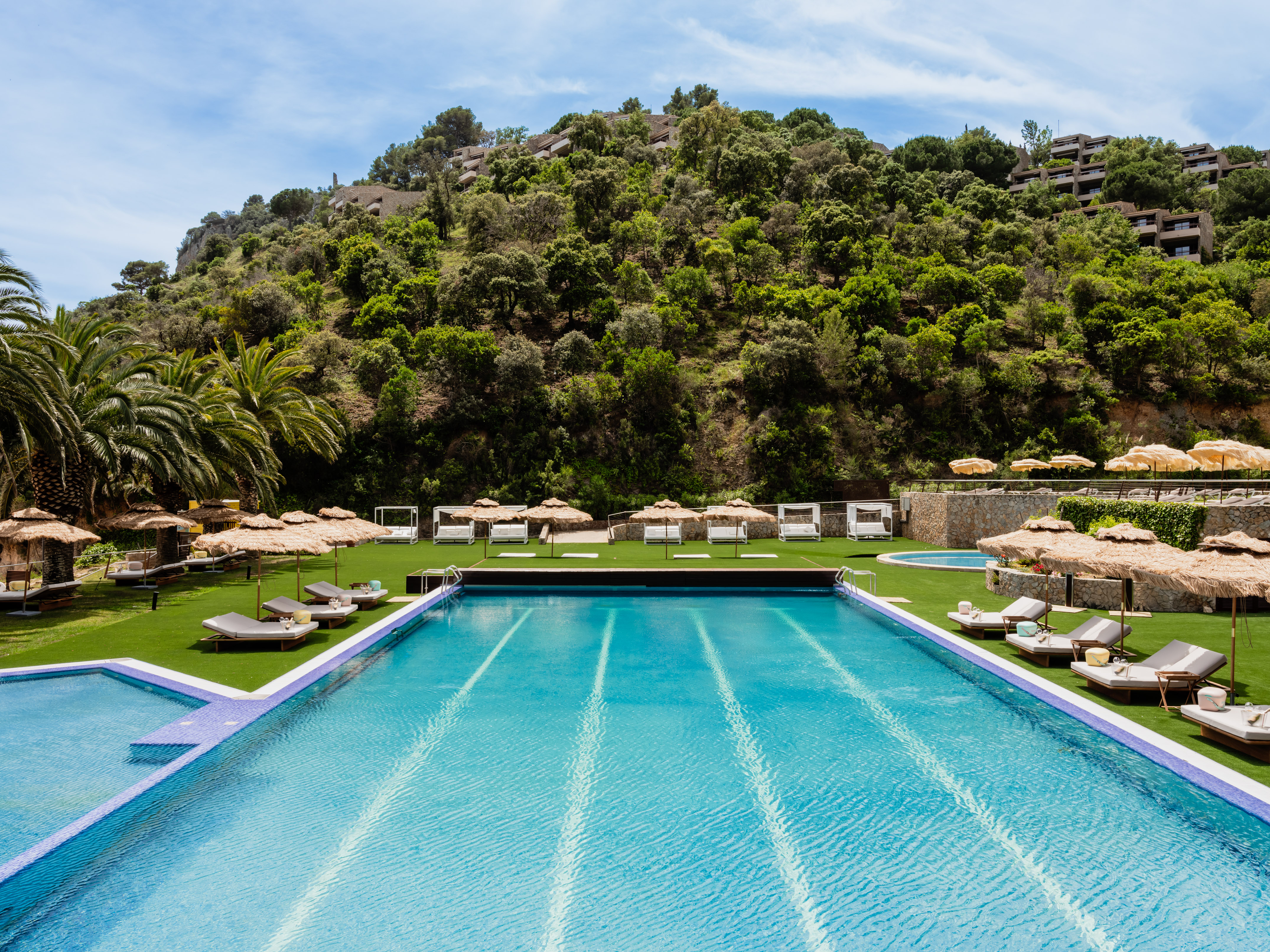 Resort swimming pool with sun loungers, straw umbrellas, and lush hillside.