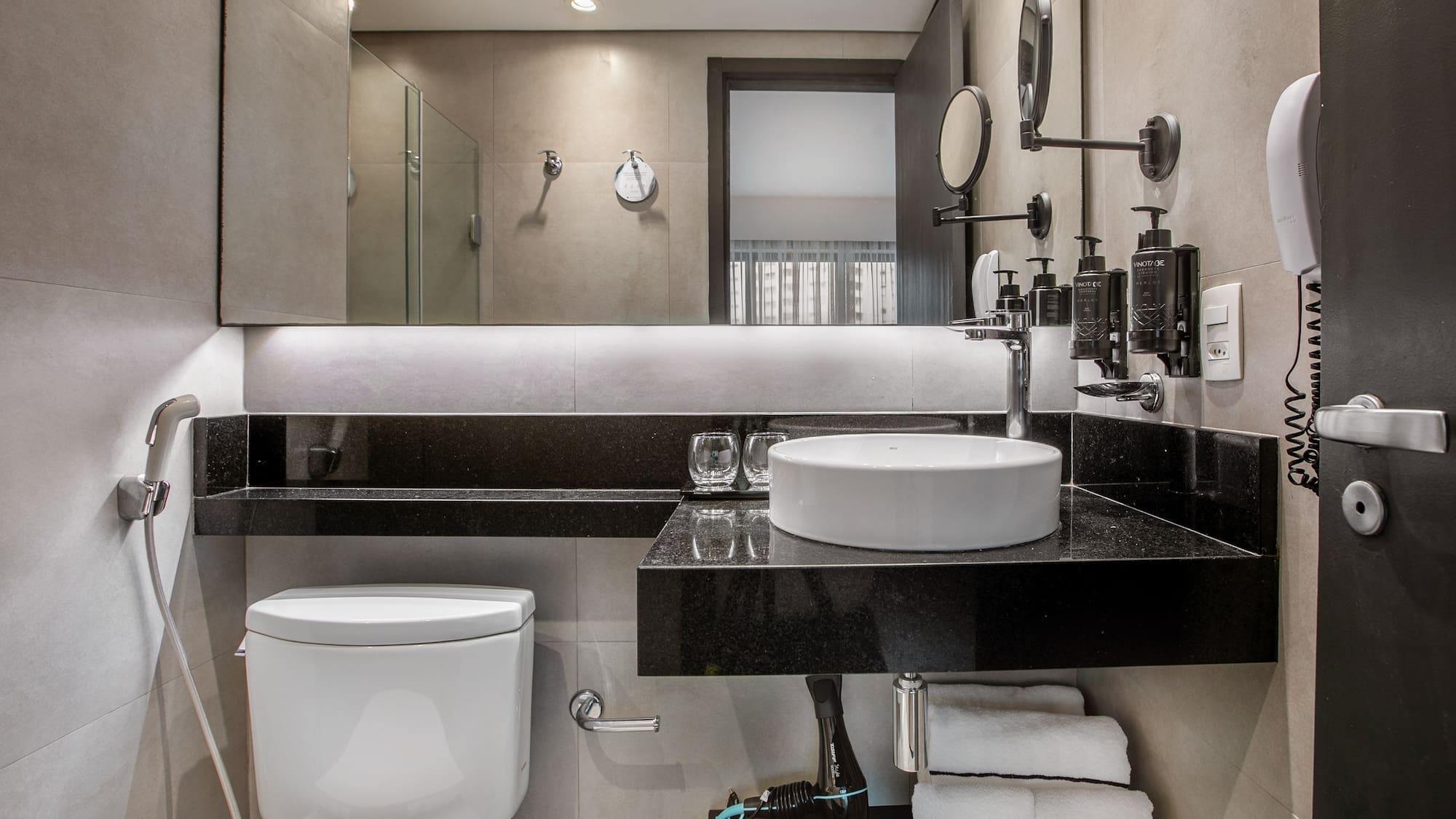 Modern hotel bathroom with a white vessel sink, black countertop, toilet, and lit mirror. - Deluxe Room Twin - Meliá Ibirapuera, São Paulo