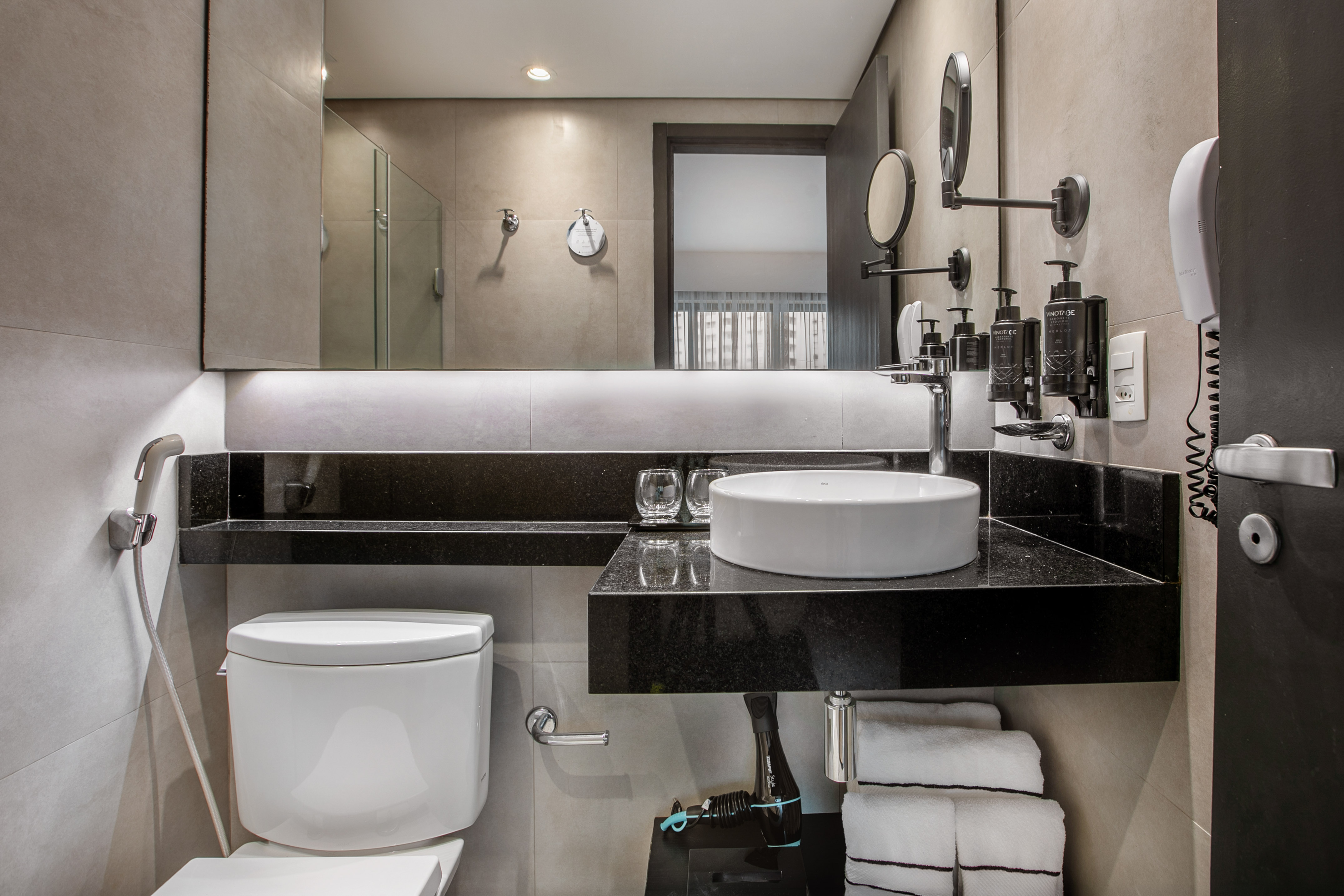 Modern hotel bathroom with a white vessel sink, black countertop, toilet, and lit mirror. - Deluxe Room Twin - Meliá Ibirapuera, São Paulo