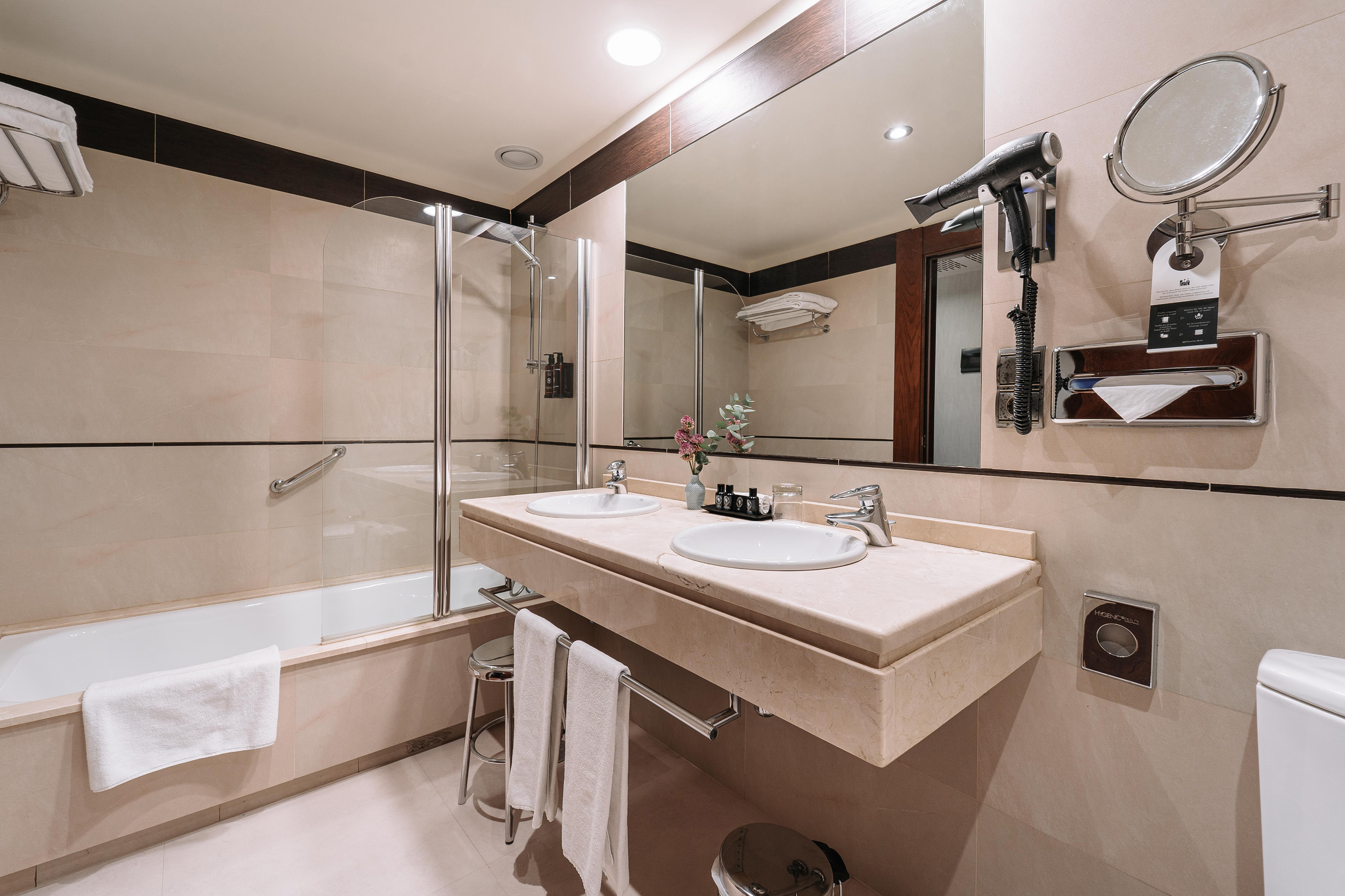 Modern hotel bathroom with double vanity, large mirror, bathtub, and hairdryer.