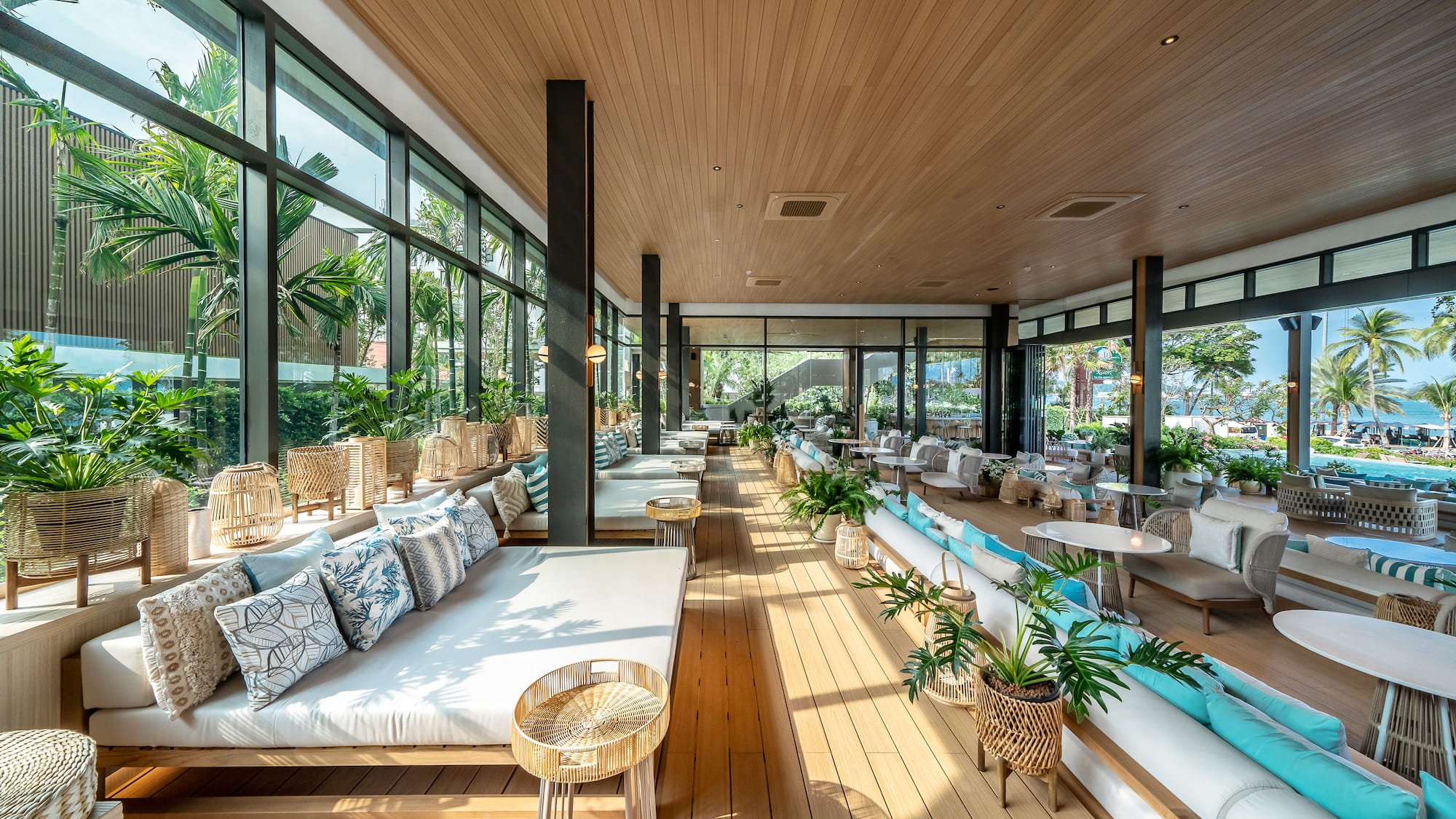 Modern resort lounge with daybeds, large windows, wood interior, and tropical view.