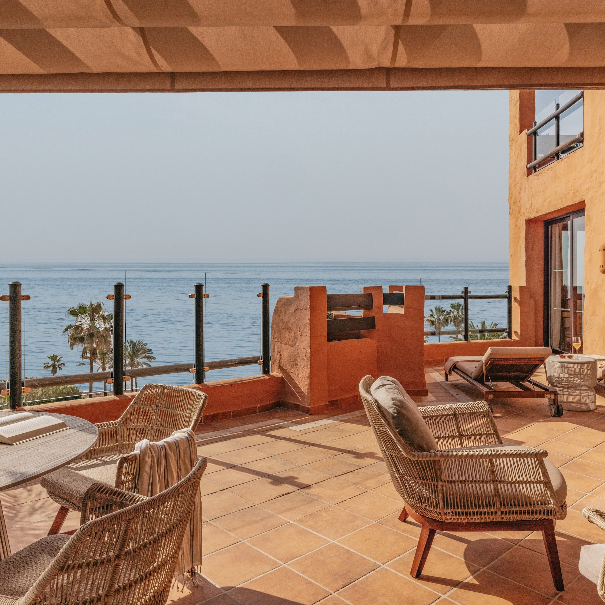 Sunny ocean view terrace with wicker chairs, wooden table, and sunbeds.
