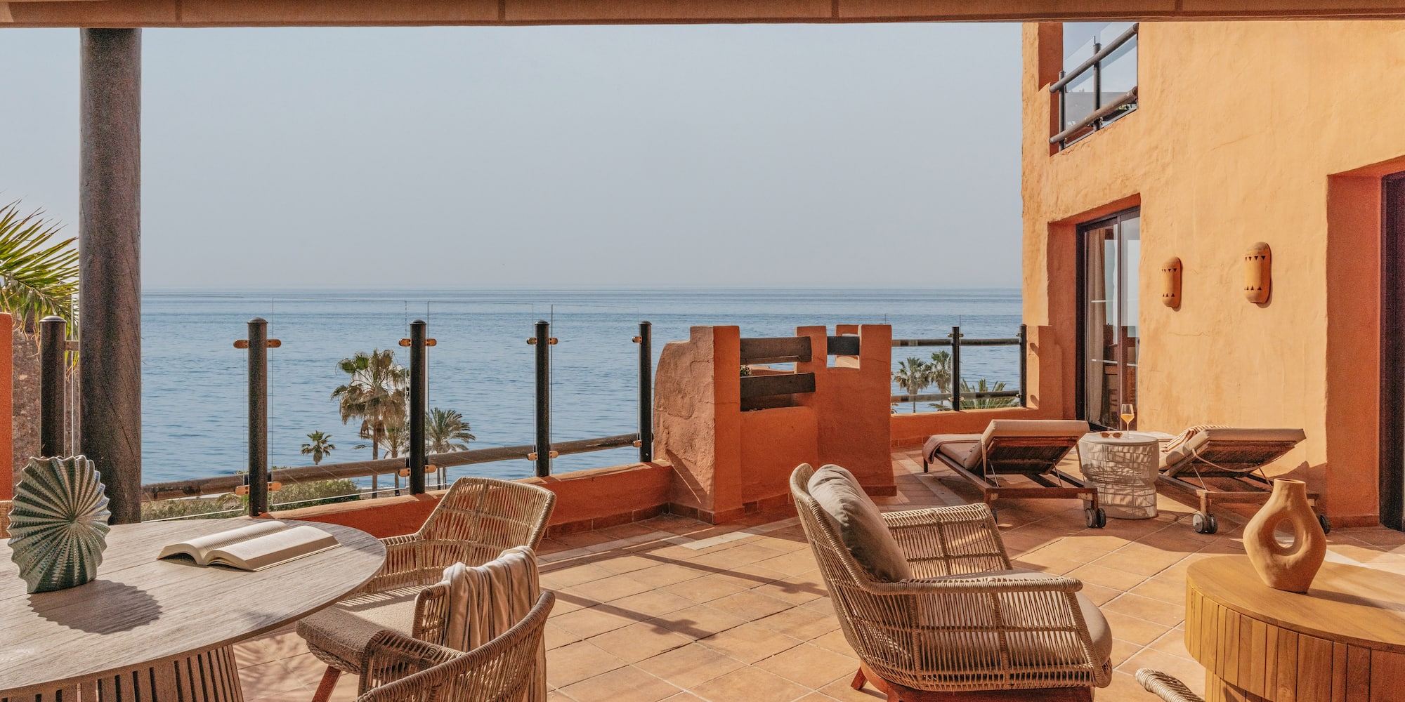 Sunny ocean view terrace with wicker chairs, wooden table, and sunbeds.