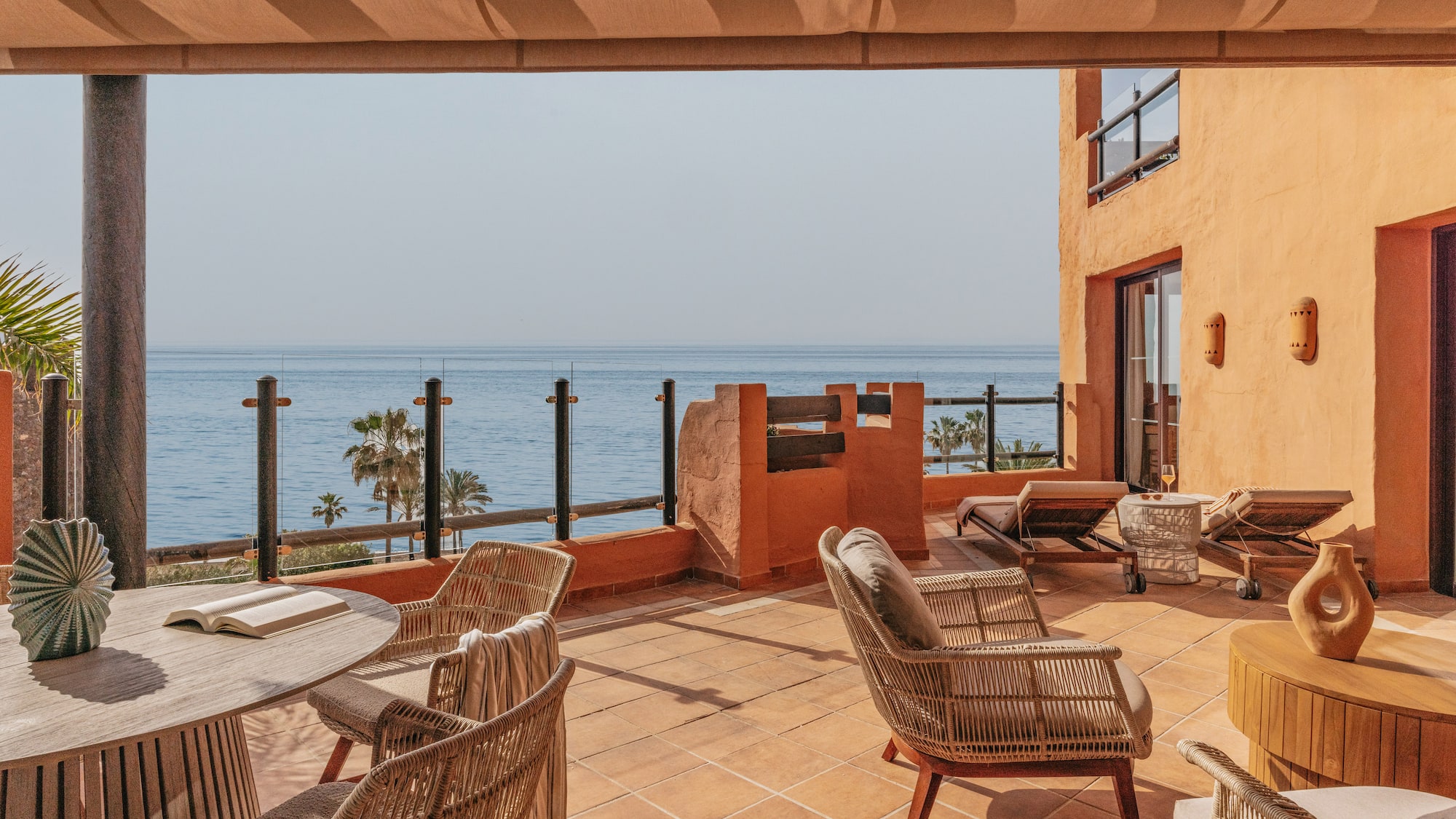 Sunny ocean view terrace with wicker chairs, wooden table, and sunbeds.