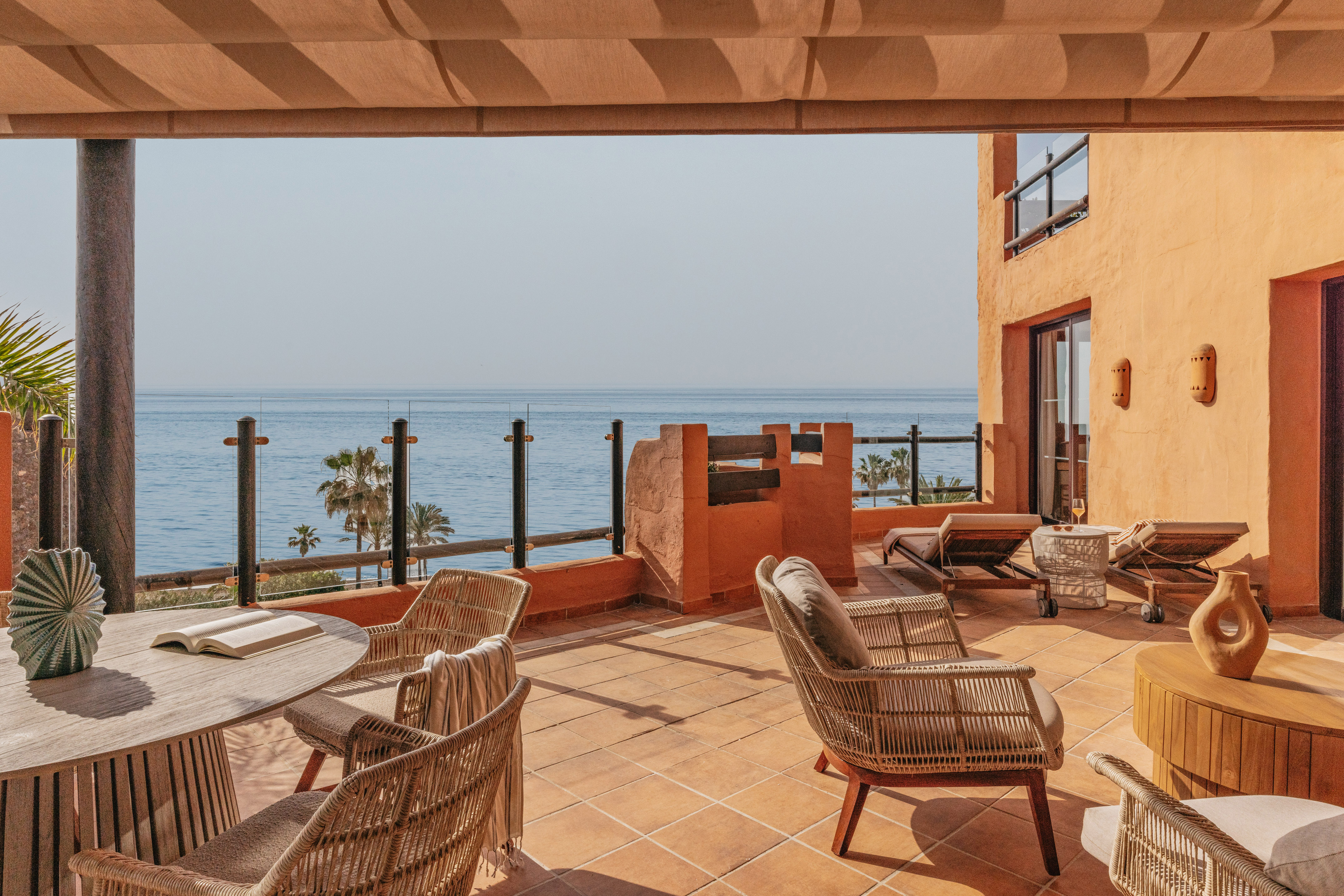 Sunny ocean view terrace with wicker chairs, wooden table, and sunbeds.