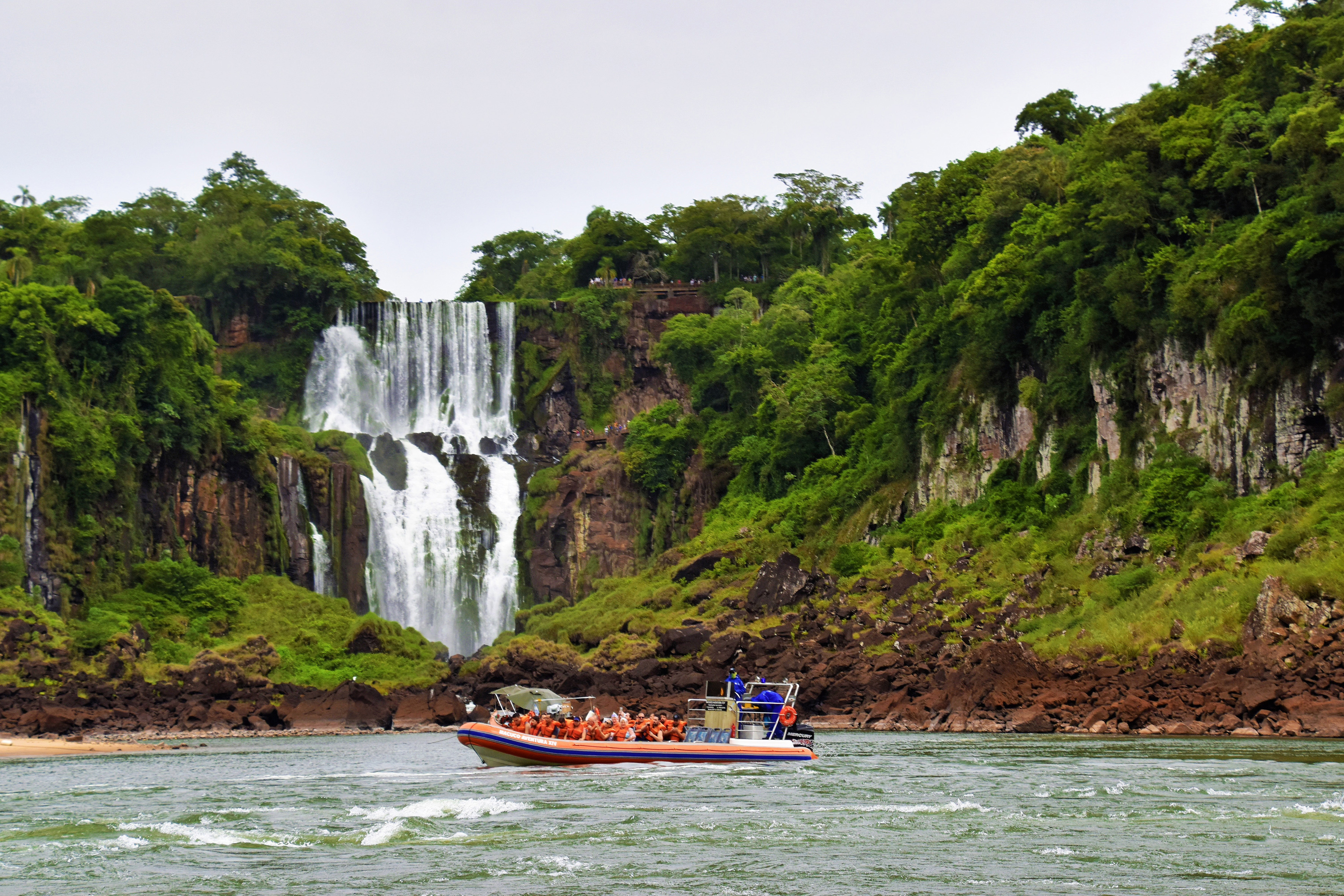 Tour boat with people at Iguazu Falls, surrounded by lush rainforest cliffs.
