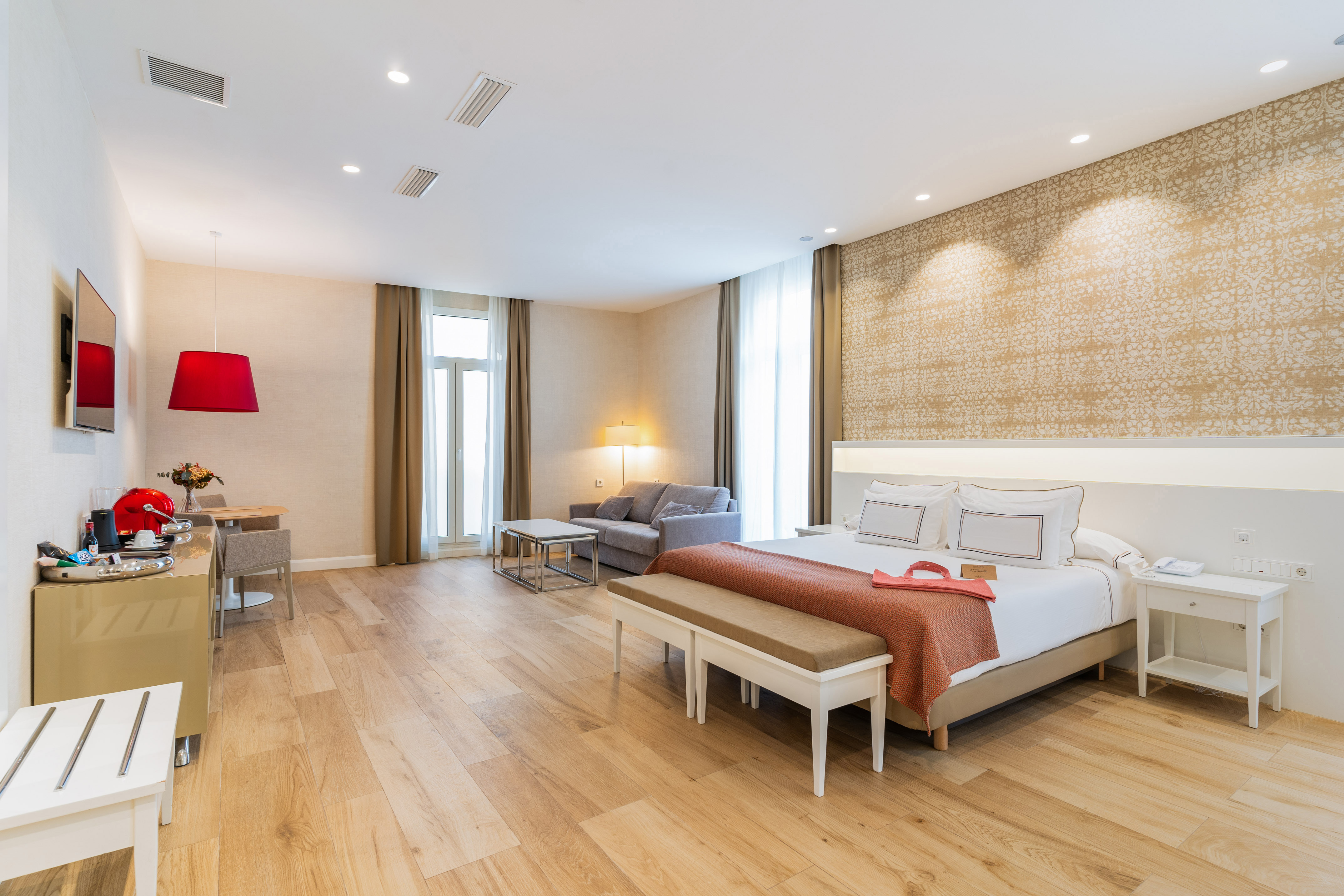 Modern hotel suite with large bed, sofa, dining area, and light wooden floor.