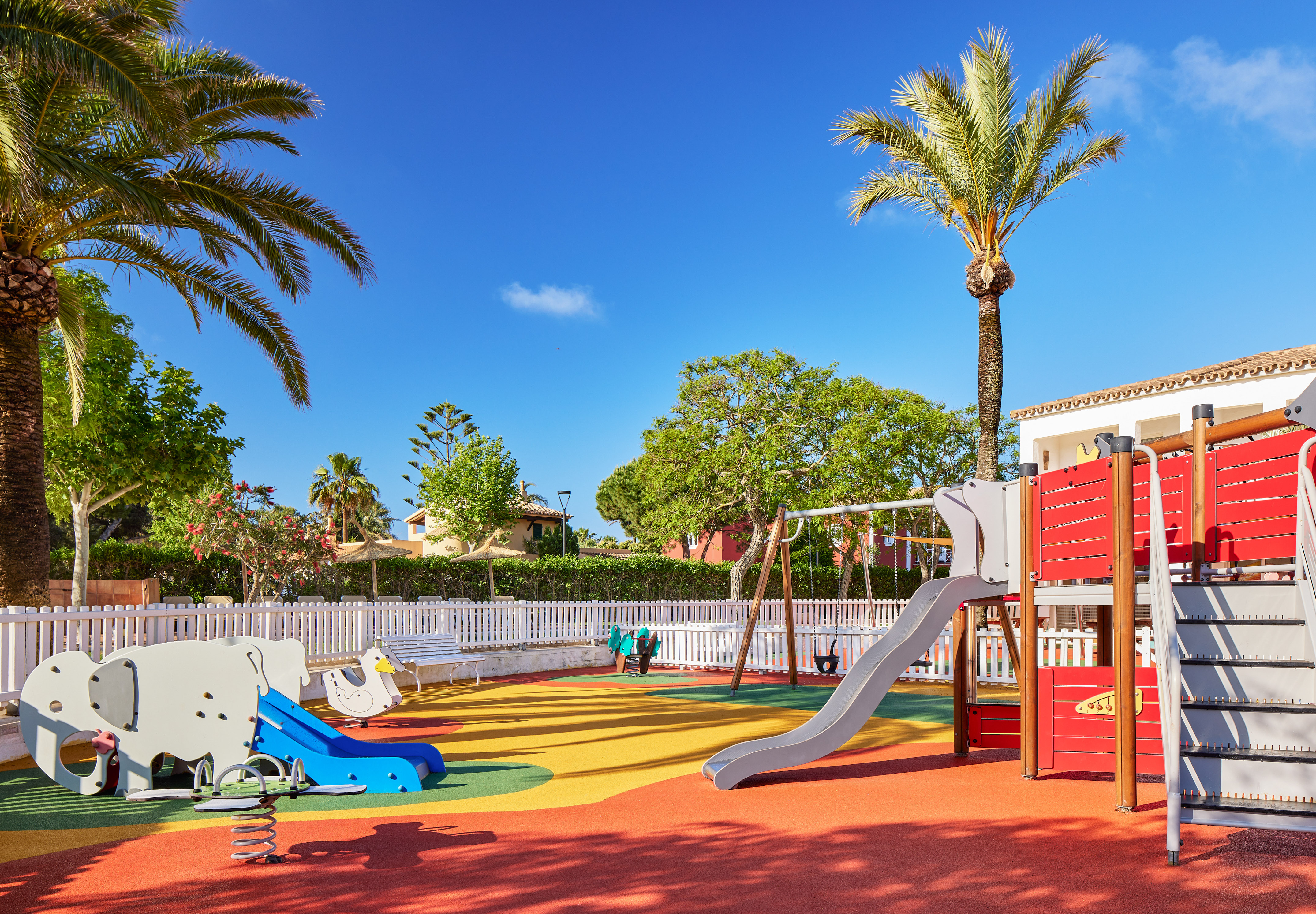 Outdoor resort playground with colorful soft ground, slide, swings, palm trees.