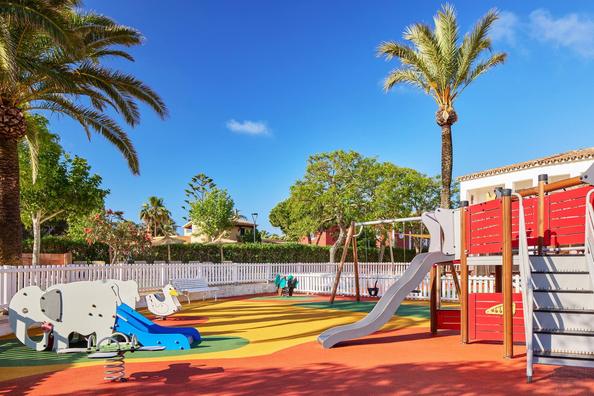 Outdoor resort playground with colorful soft ground, slide, swings, palm trees.