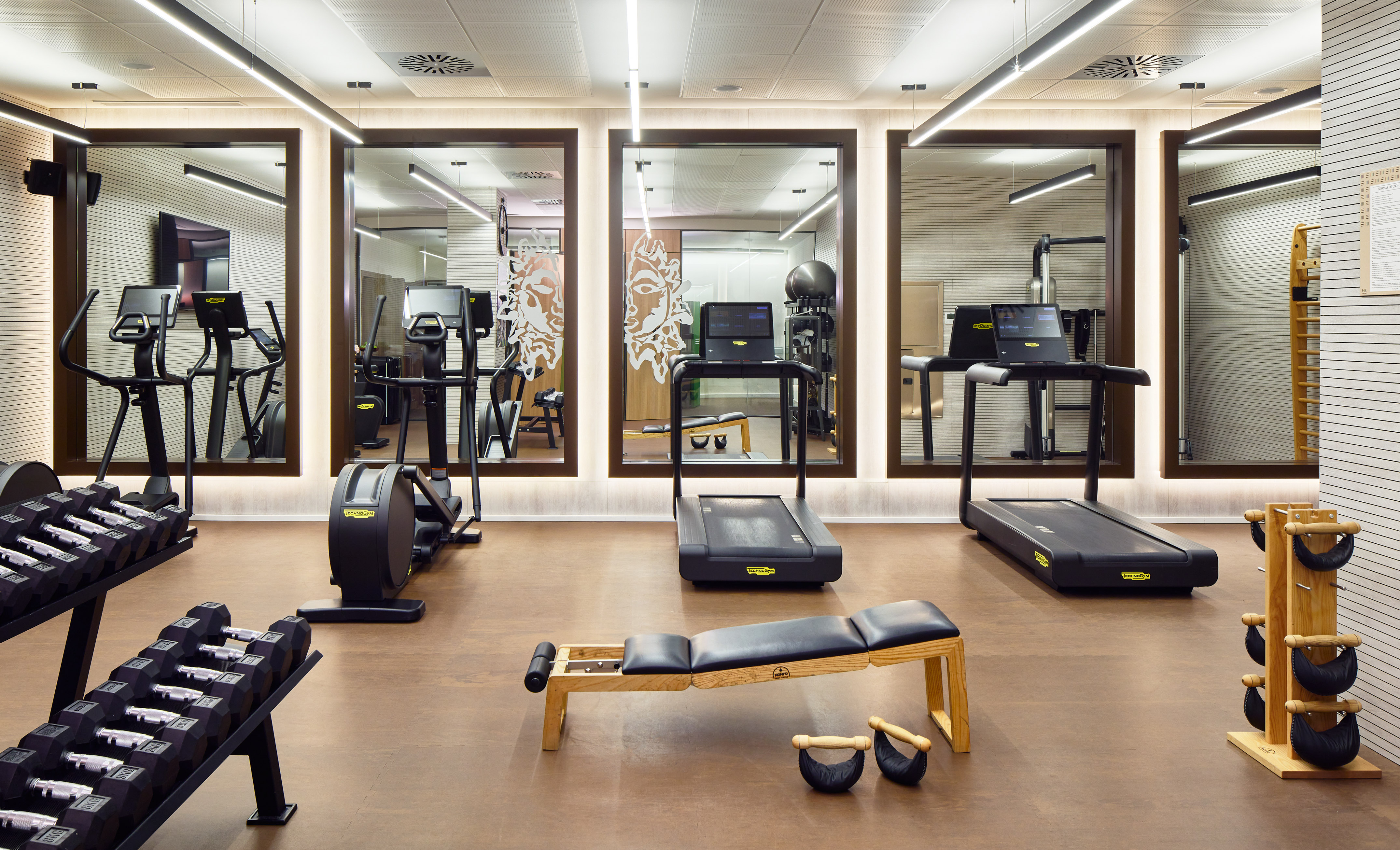 Modern hotel gym with treadmills, ellipticals, dumbbells, and workout bench.