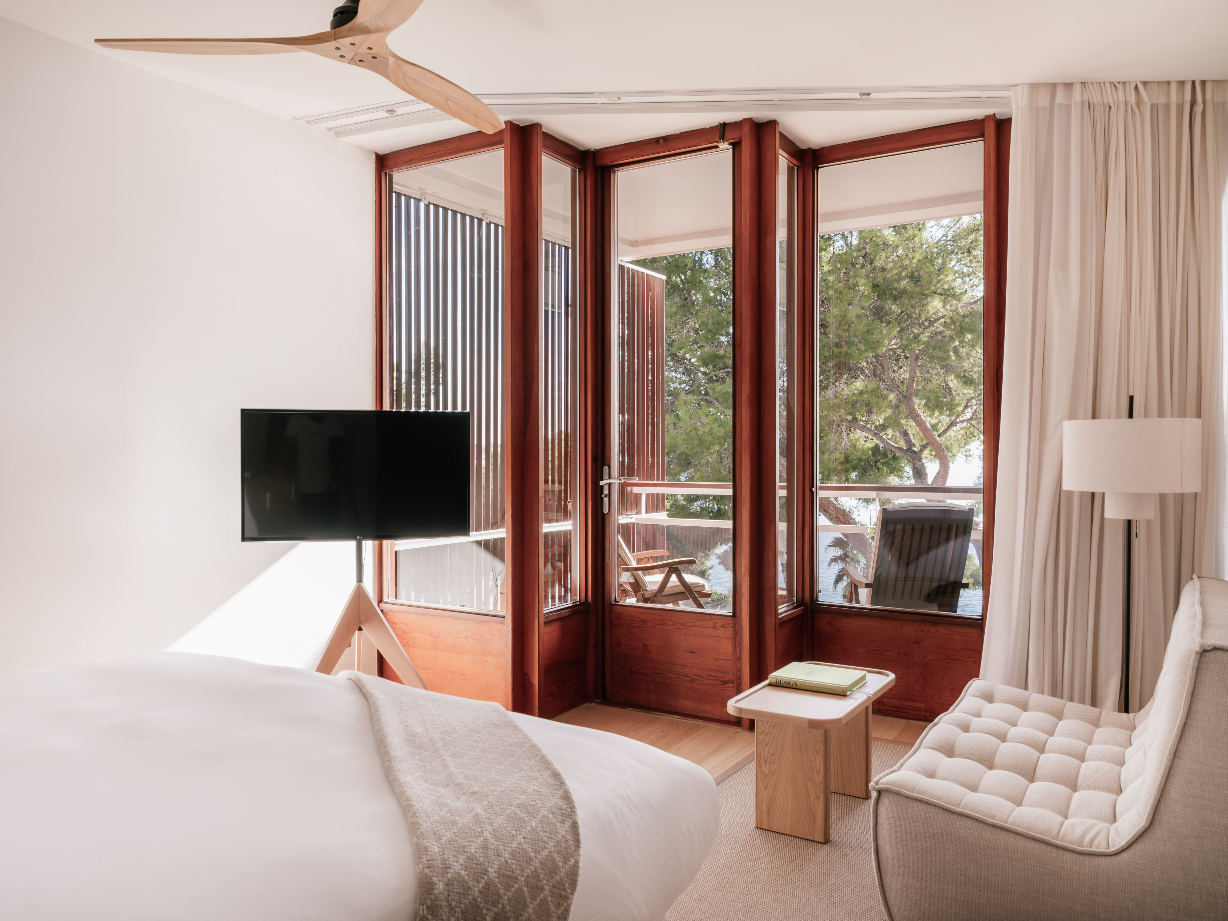 Modern hotel bedroom with large windows, balcony, and a serene tree view.