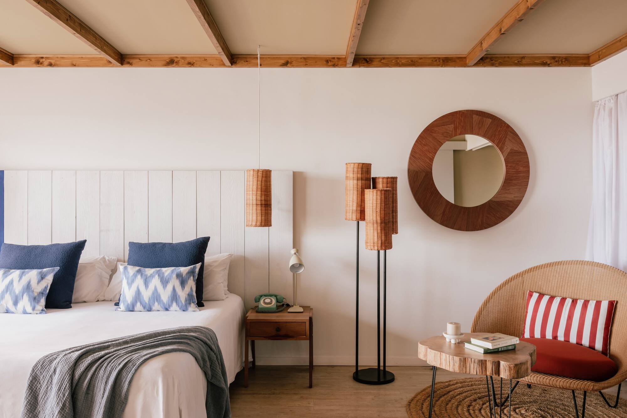Cozy bedroom with white bed, blue pillows, wood ceiling, wicker chair, rattan lamps.