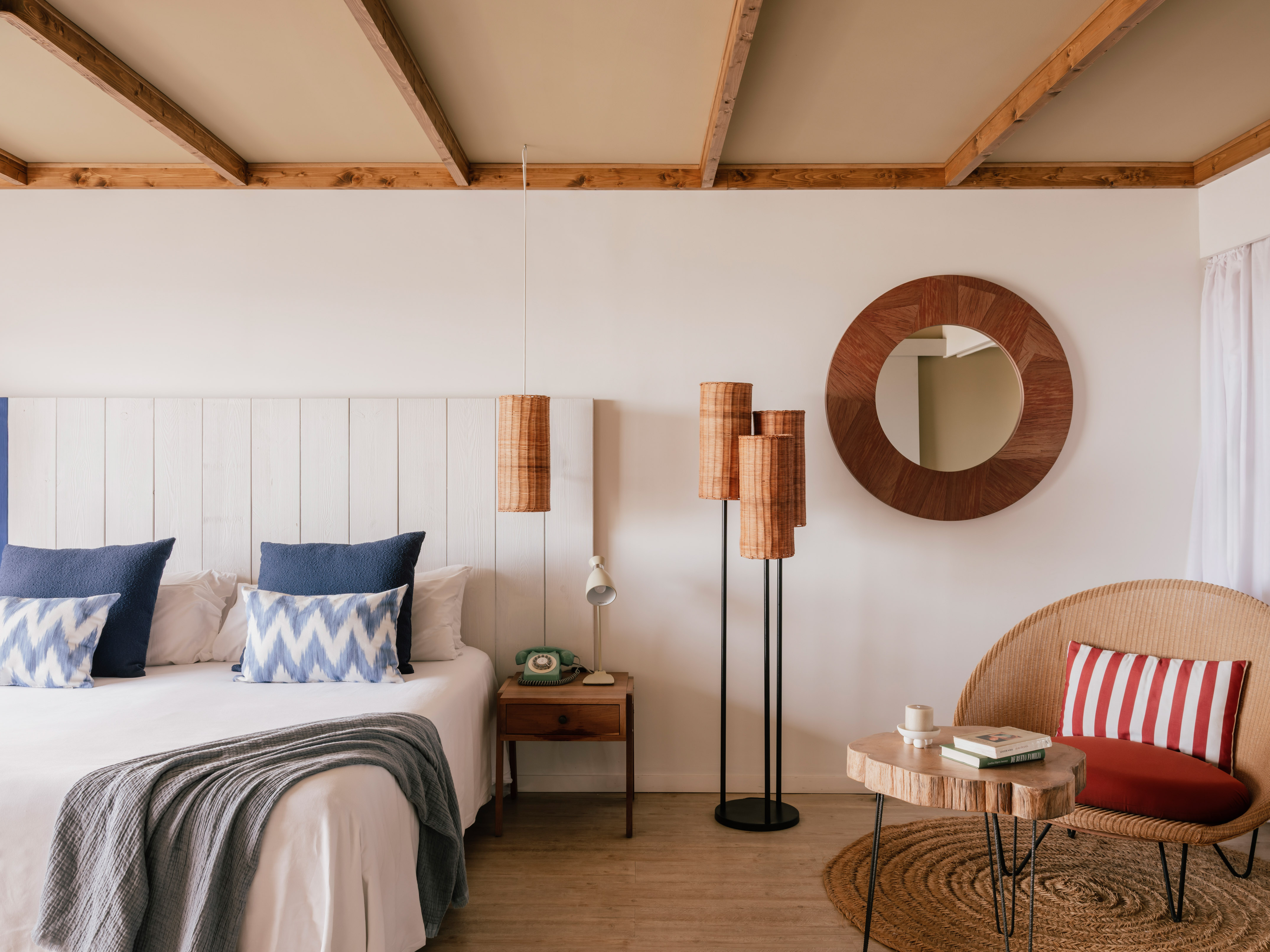 Cozy bedroom with white bed, blue pillows, wood ceiling, wicker chair, rattan lamps.