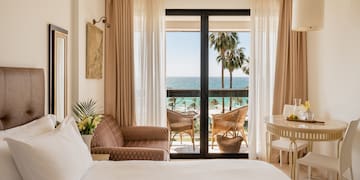 a room with a bed and chairs and a table and chairs and a balcony with a view of the ocean