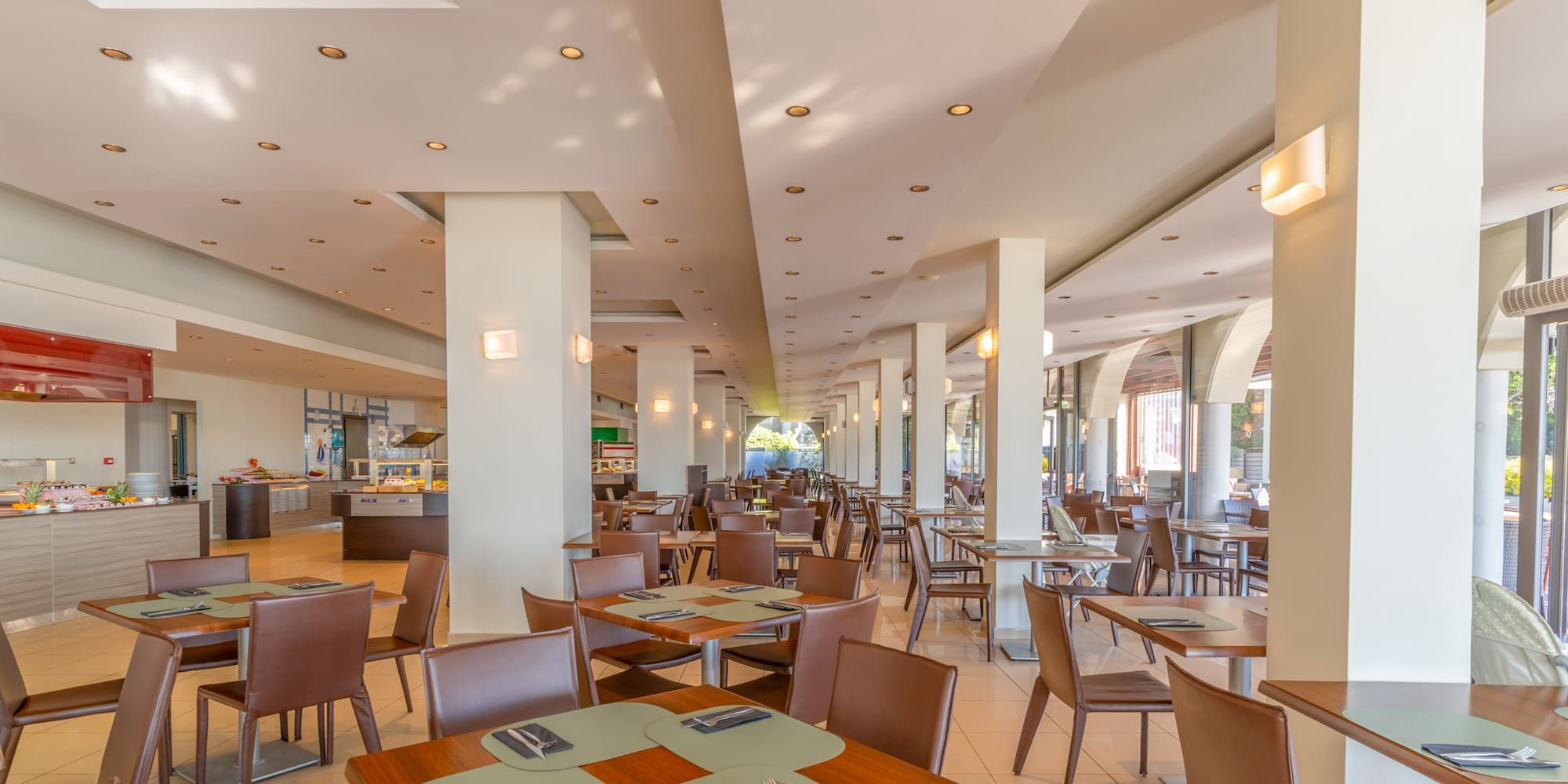 Spacious hotel dining hall with modern tables, brown chairs, and buffet area.
