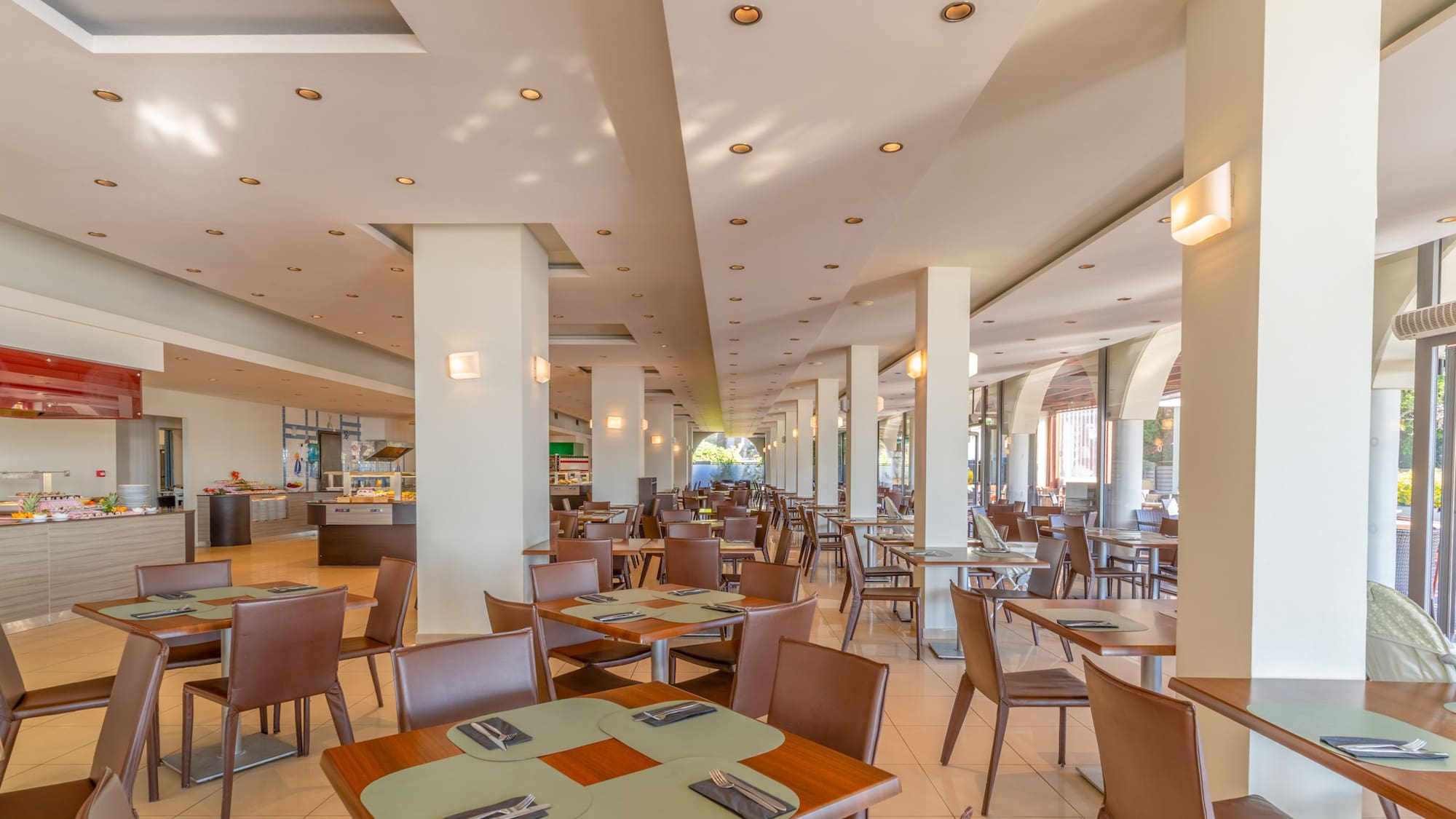 Spacious hotel dining hall with modern tables, brown chairs, and buffet area.