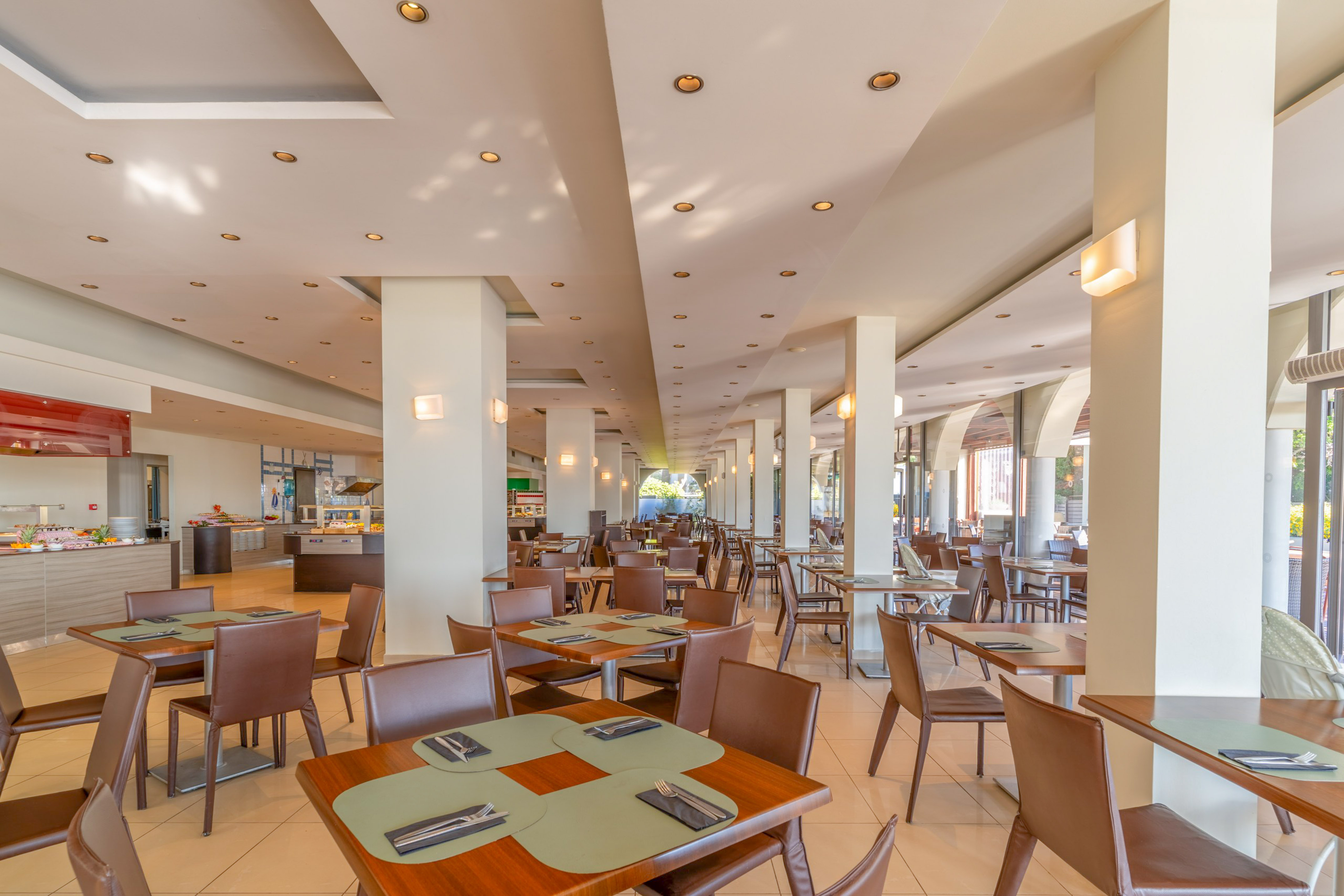 Spacious hotel dining hall with modern tables, brown chairs, and buffet area.