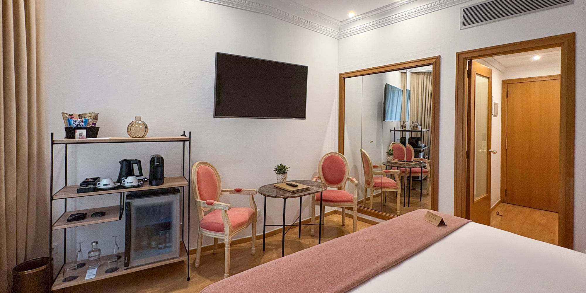 Modern hotel room with bed, minibar, wall-mounted TV, pink chairs, and large mirror.