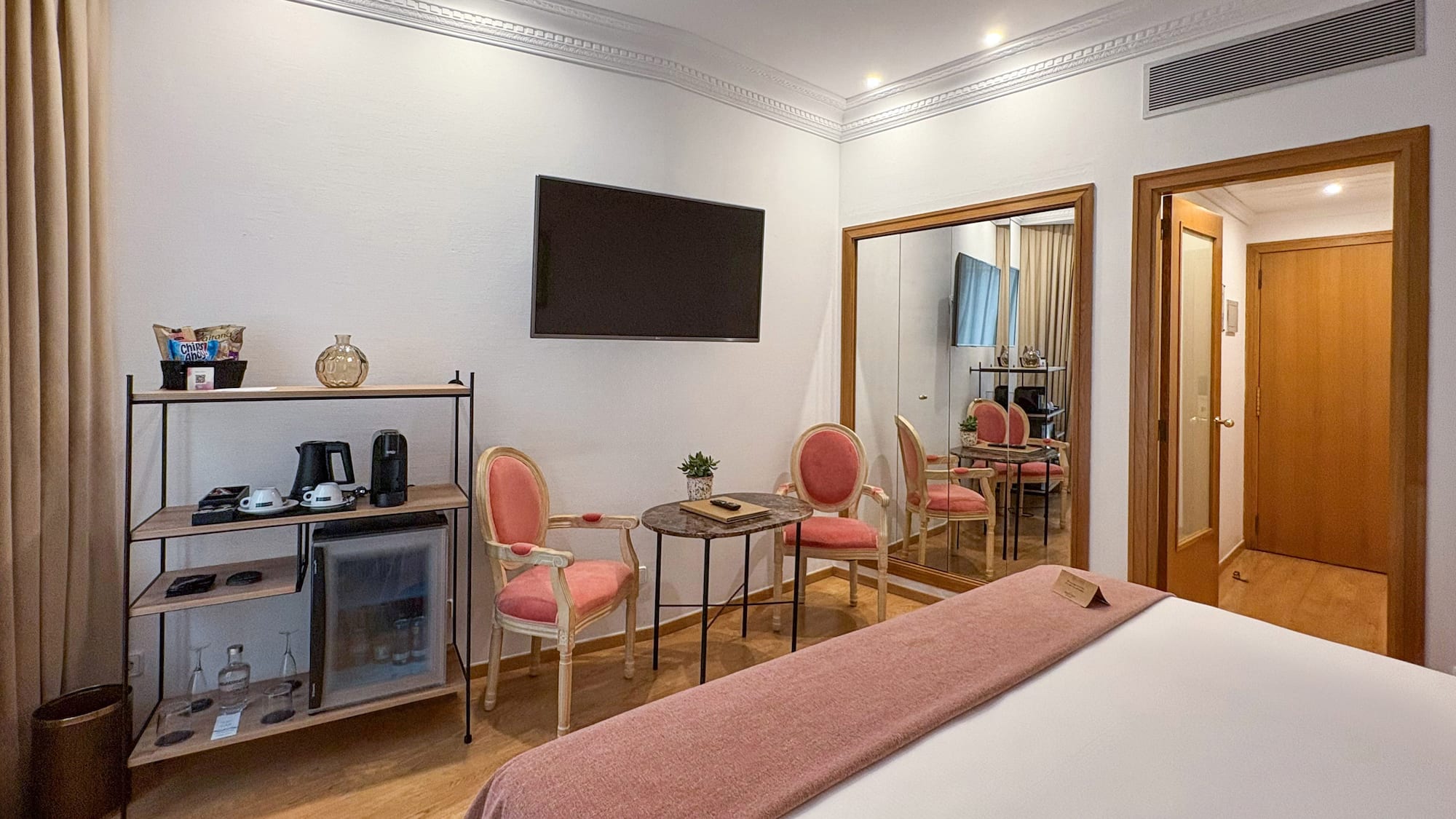Modern hotel room with bed, minibar, wall-mounted TV, pink chairs, and large mirror.